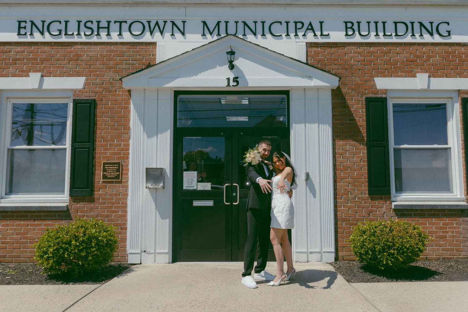 Couple embracing outside Englishtown Municipal Building, NJ, celebrating their courthouse wedding on a sunny day.