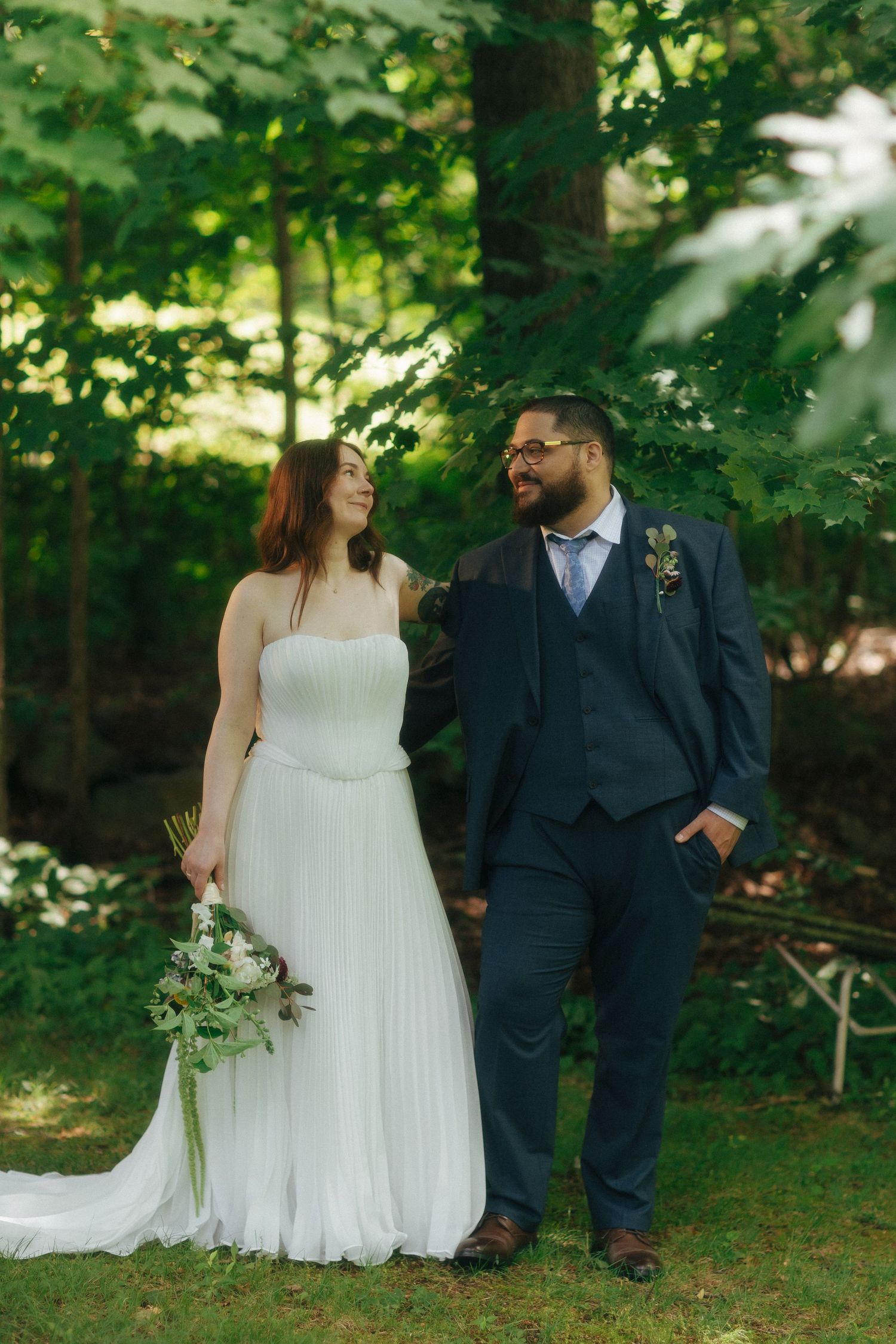 Bride in white pleated gown holding bouquet stands with groom in navy suit among lush green trees.