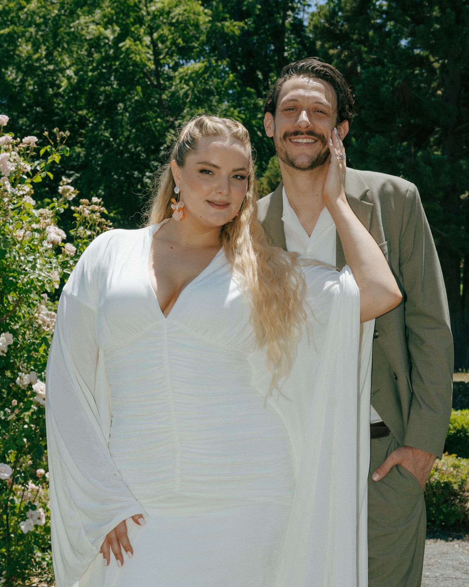 A couple in wedding attire poses outdoors, woman in white gown, man in olive suit, surrounded by lush greenery.