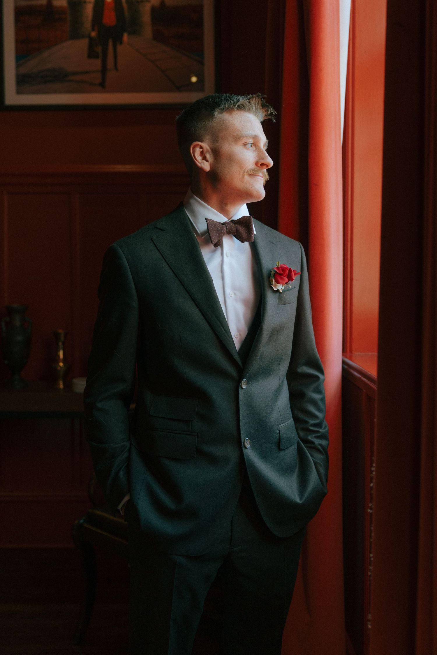 Groom in dark green tuxedo with red boutonniere and bow tie stands by red wall looking out window.