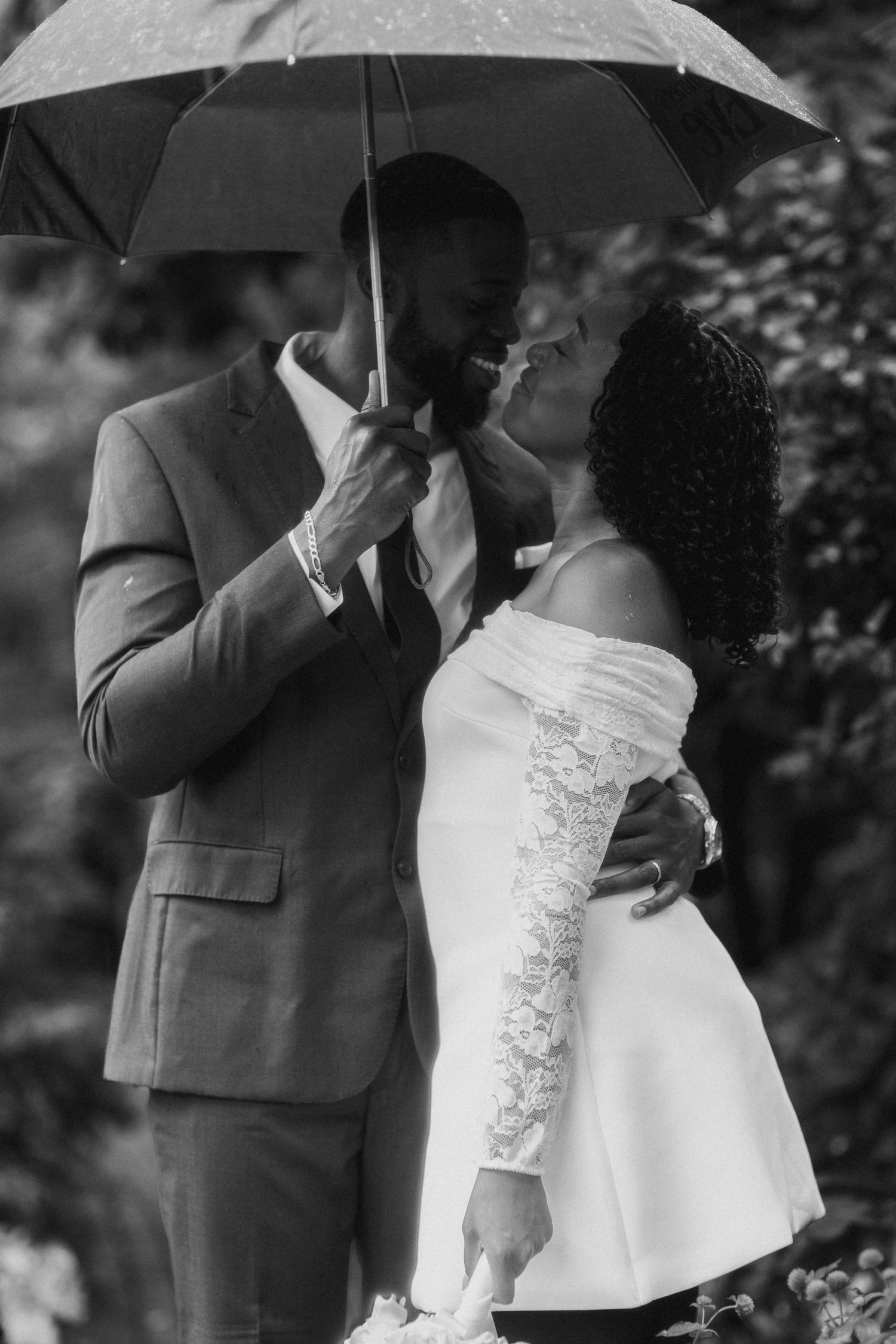 Black and white photo of newlyweds sharing a romantic moment under umbrella in the rain.