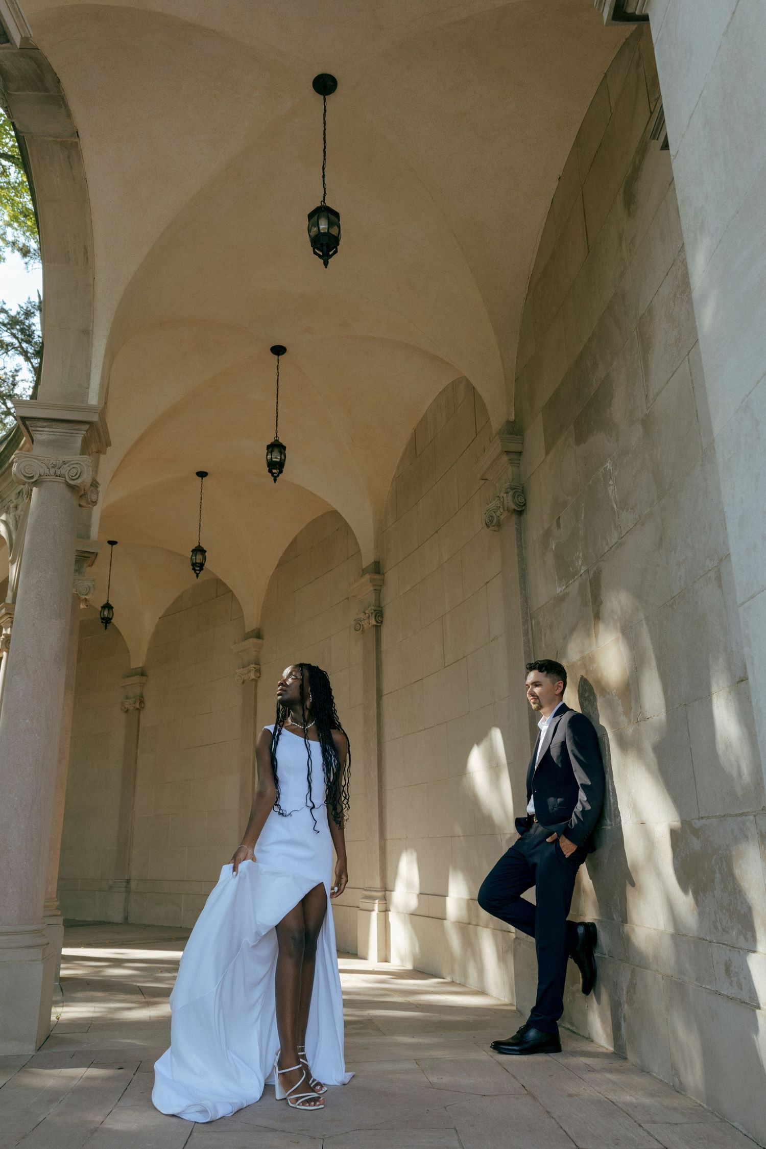 Bride in white gown and groom in dark suit pose under elegant stone arched corridor with hanging lanterns.