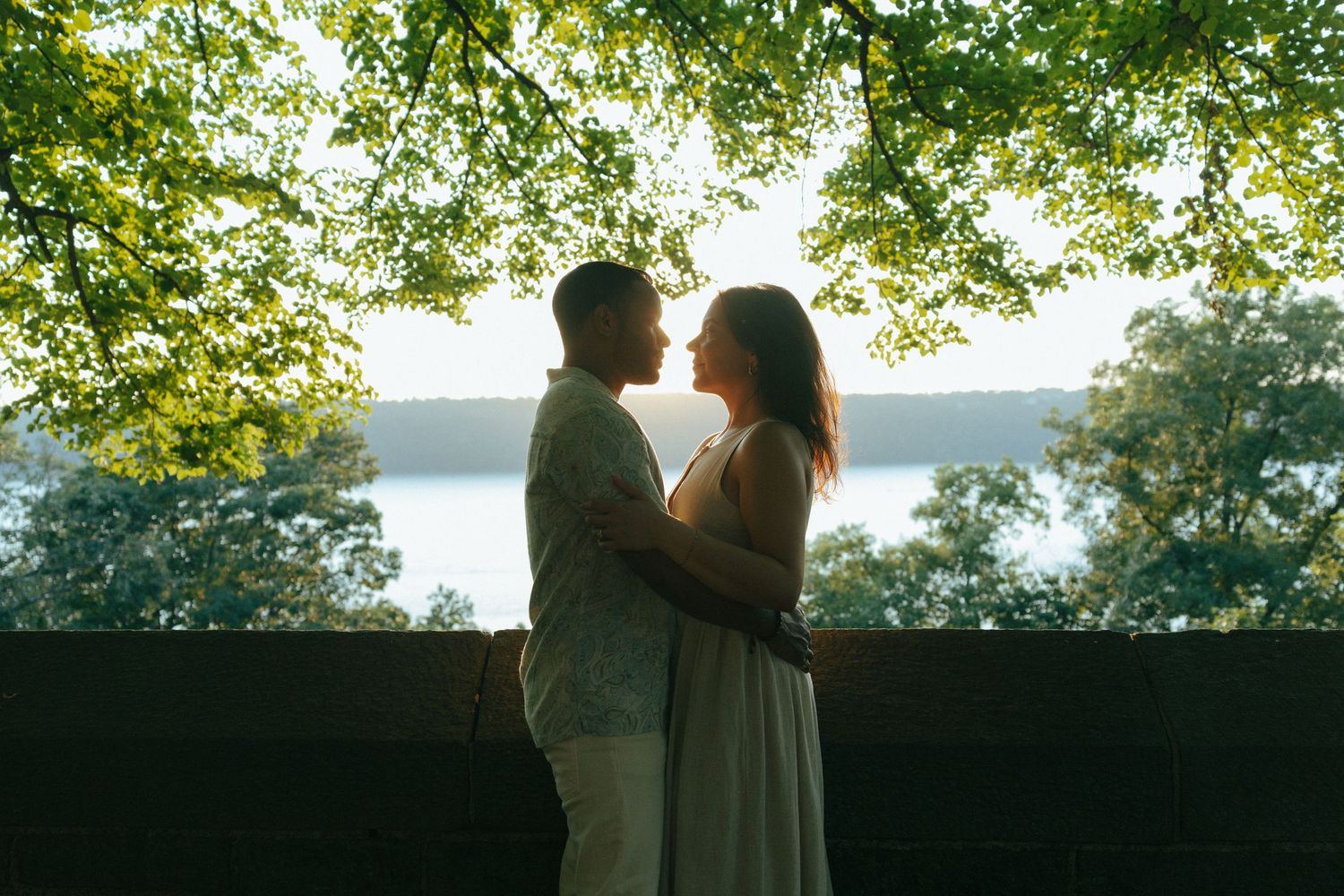 Romantic couple embracing at sunset under lush green trees with a serene lake view in the background.