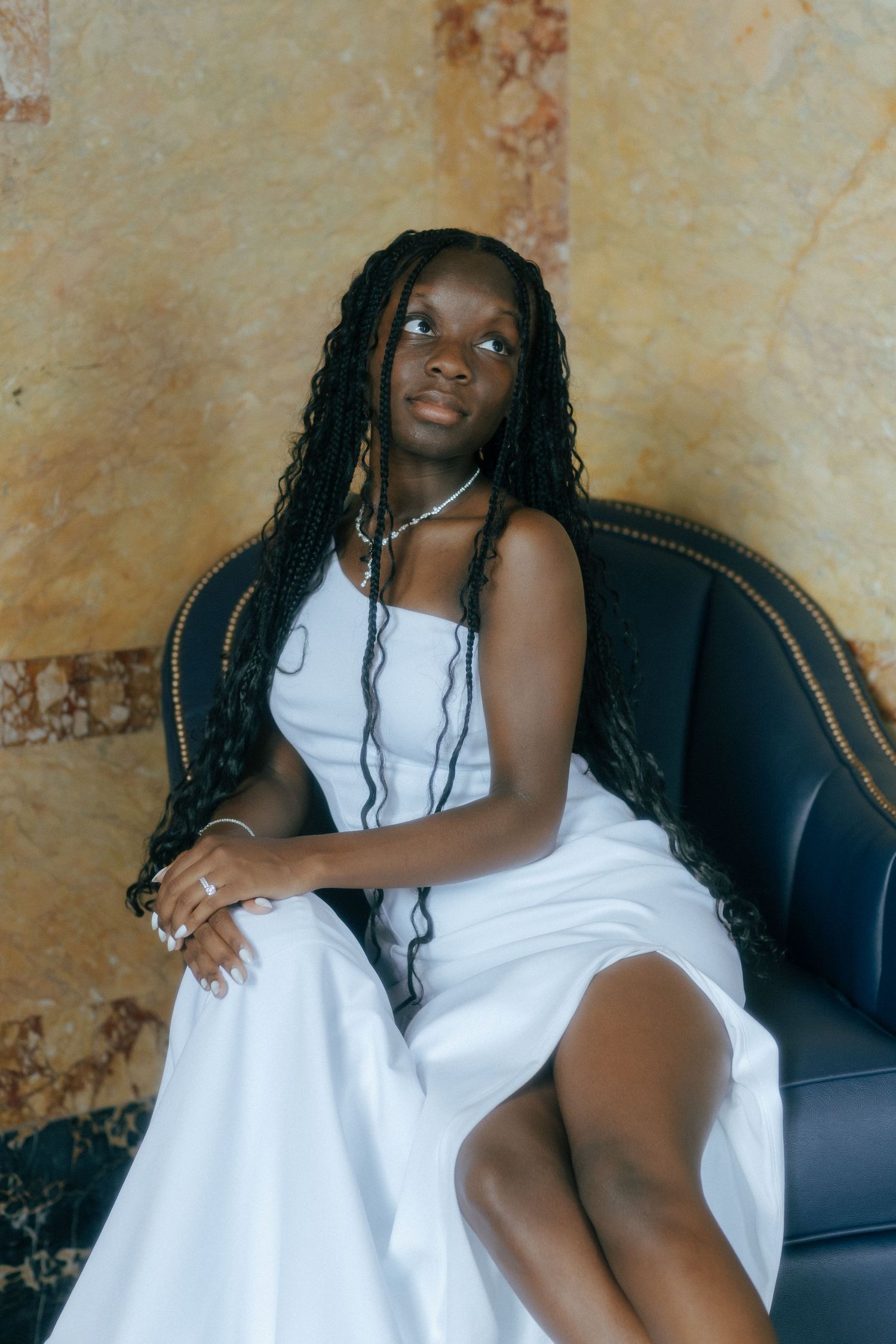 Young Black woman with long braids in white dress seated elegantly on dark leather chair against marble wall.