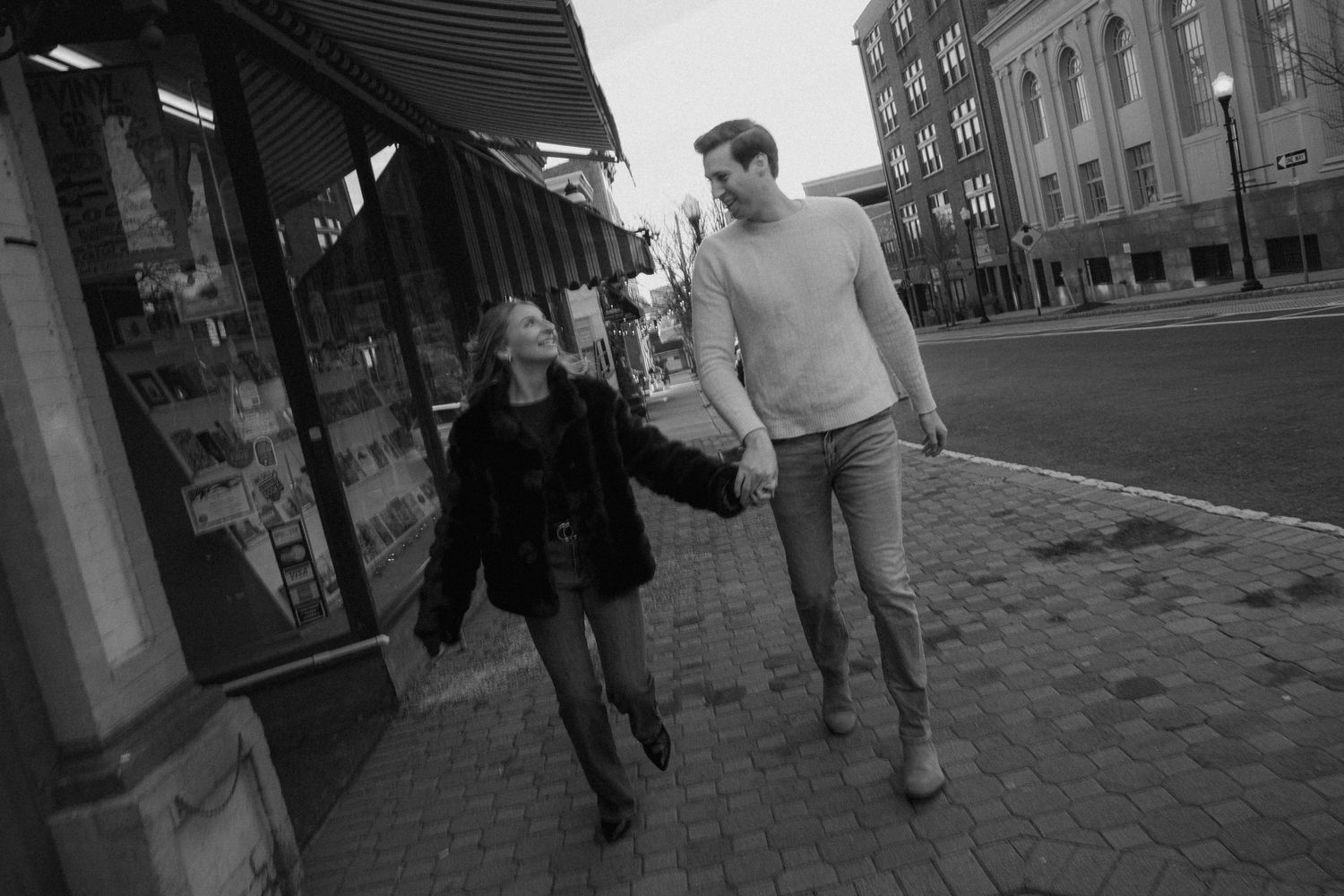 Black and white photo of a couple holding hands and strolling past a storefront on a city sidewalk.