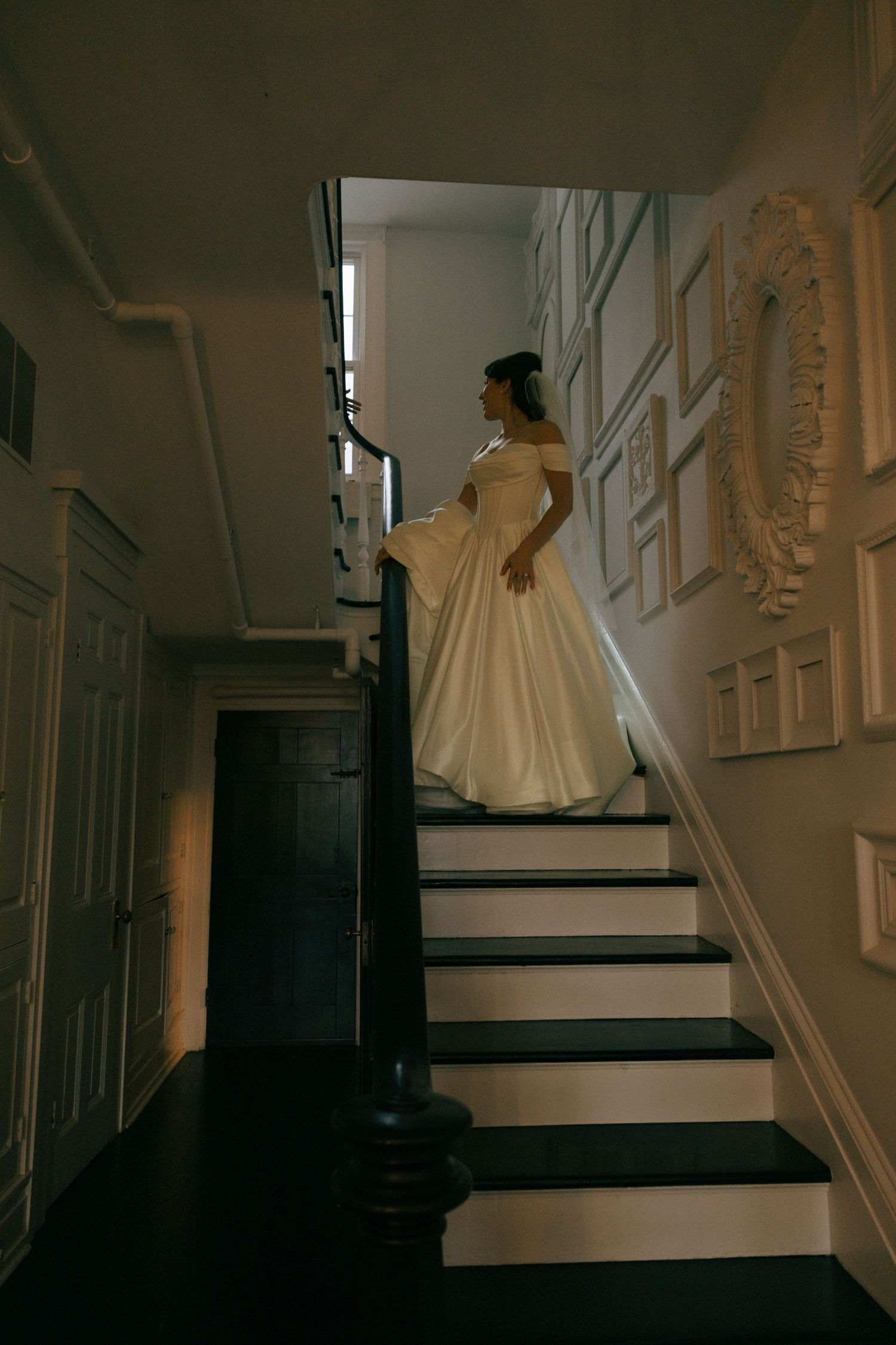 Bride in elegant white ball gown standing on dark staircase in vintage mansion with ornate wall decor.