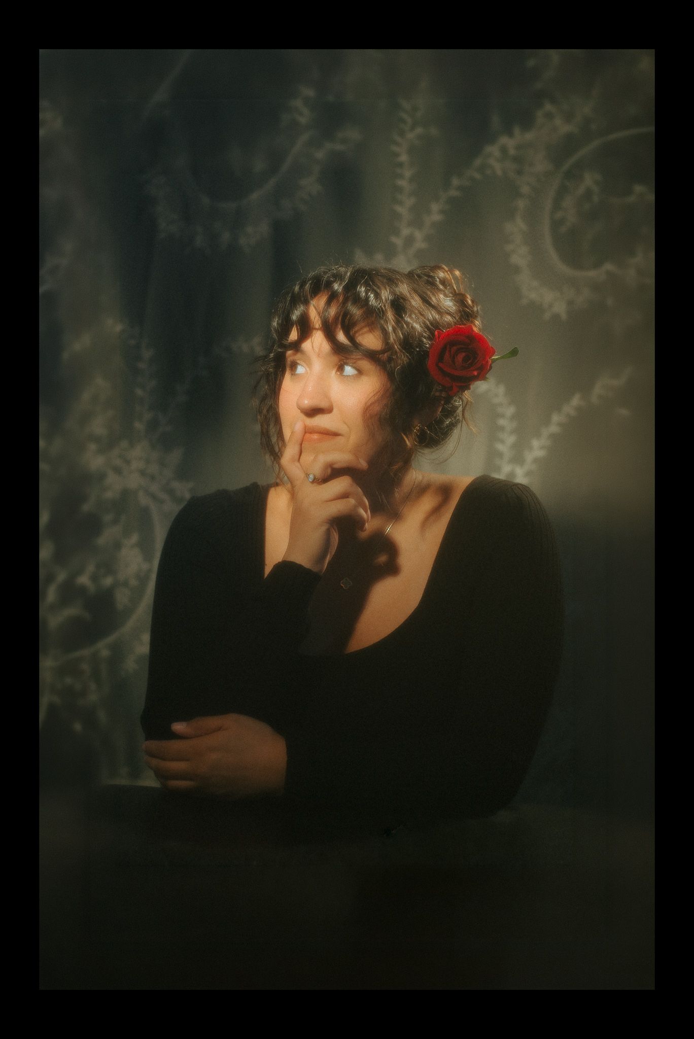 A pensive woman with curly hair adorned with a red rose, wearing a black top, posing thoughtfully against a floral backdrop.