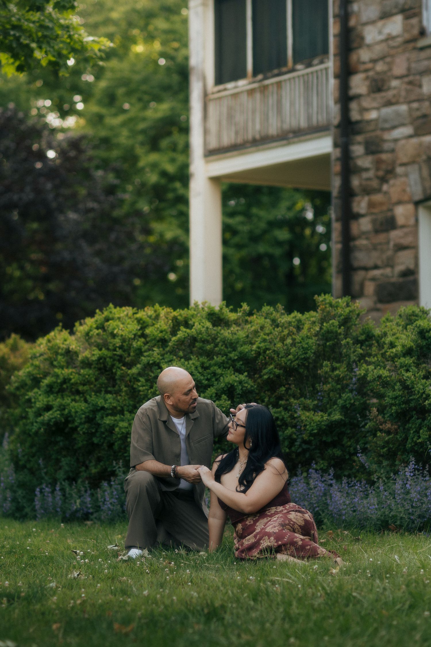 Couple sitting together on lush green grass in front of a stone building, sharing a tender moment outdoors.