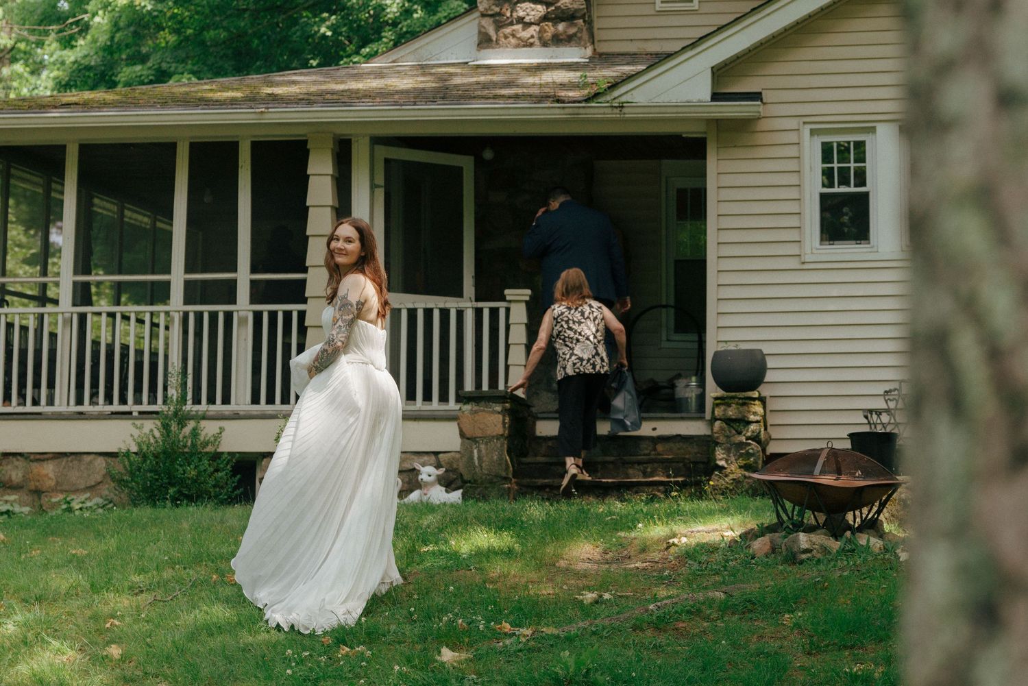 Bride in white gown twirling in yard outside rustic farmhouse with guests on porch.