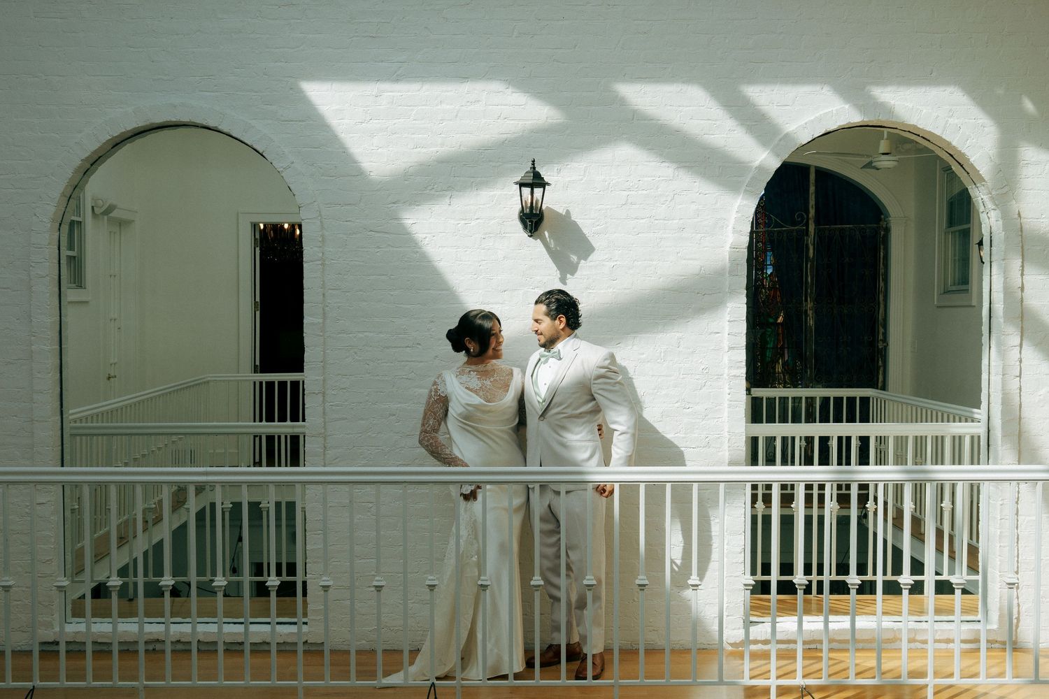 Bride and groom share a romantic moment on a white balcony with arched windows at their elegant wedding venue.