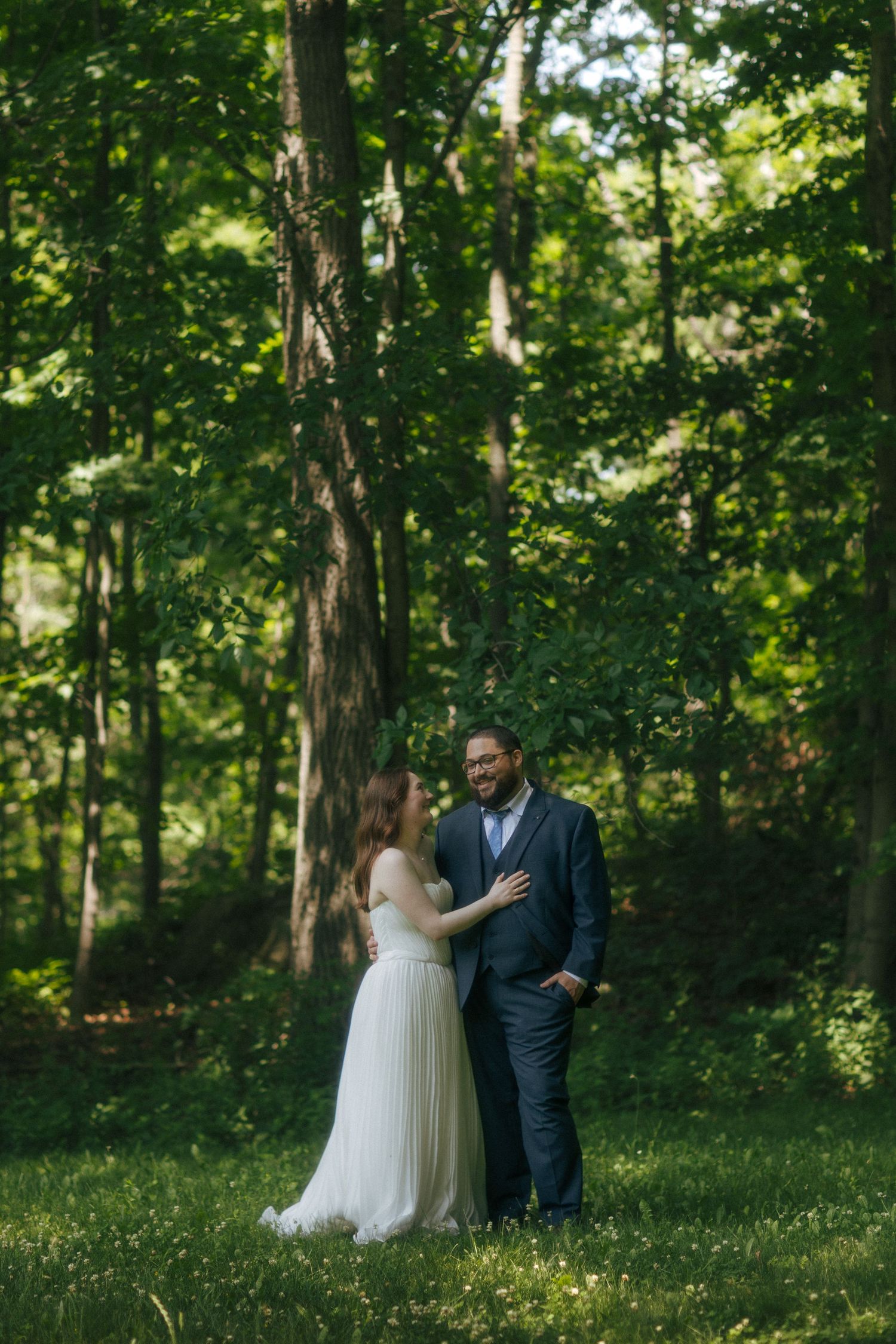 Bride and groom share a romantic moment in a lush green forest, bride in white gown, groom in navy suit.