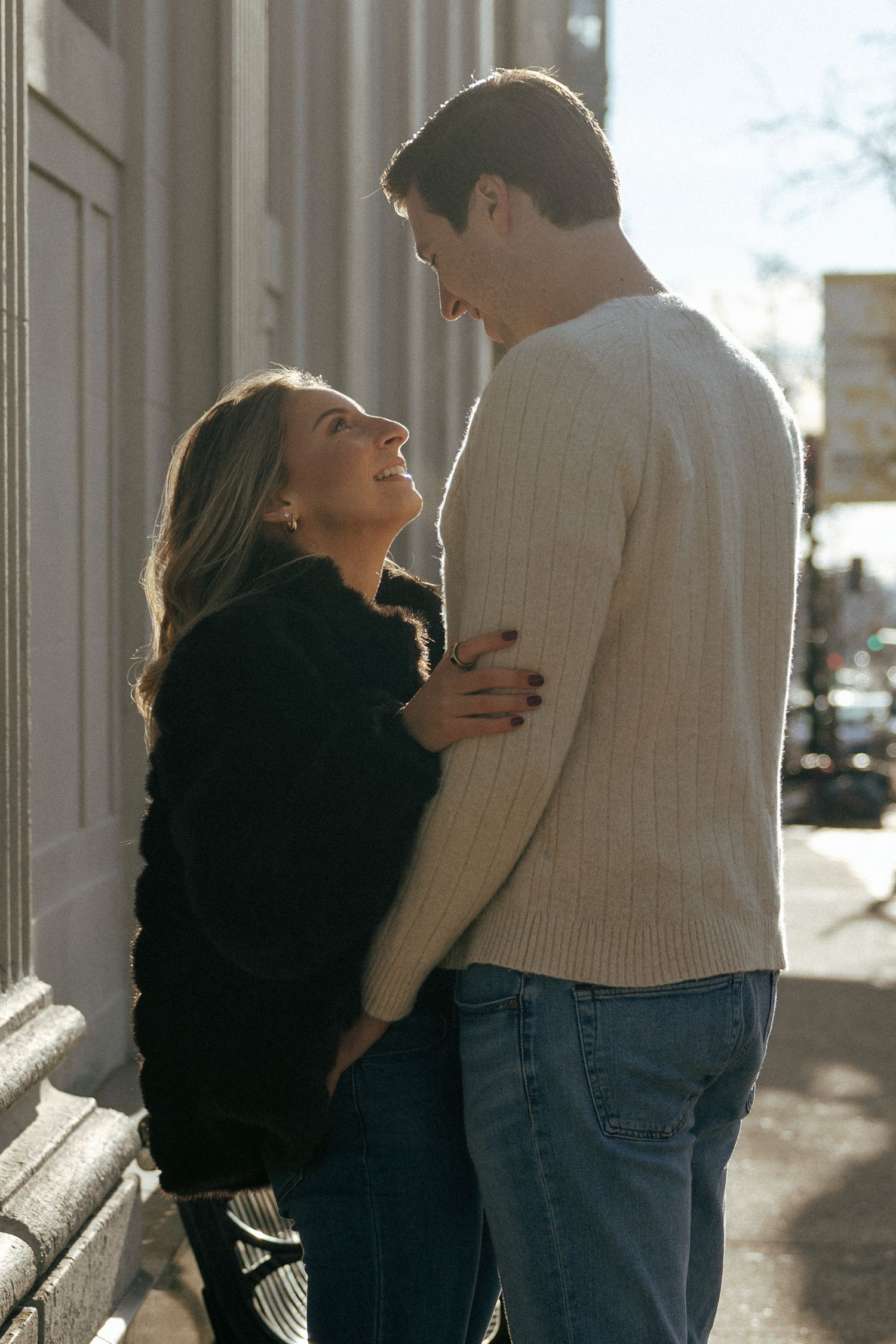 Romantic couple embracing and gazing at each other on sunny urban sidewalk, woman in black jacket, man in cream sweater.