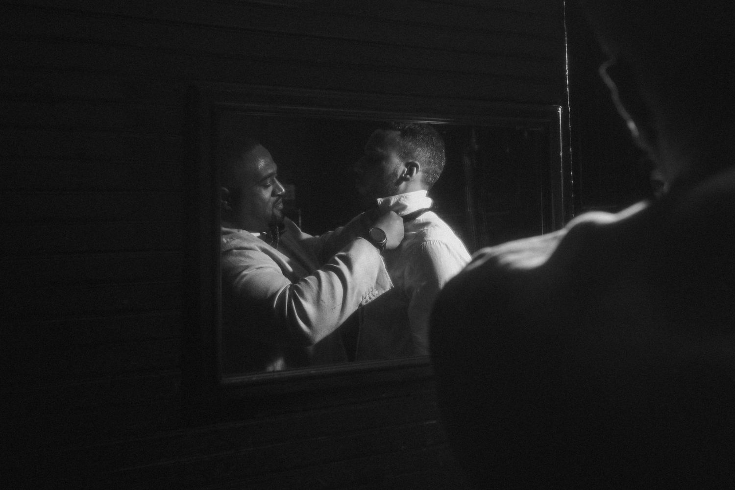 Black and white photo of groom adjusting tie in mirror reflection during wedding preparations.