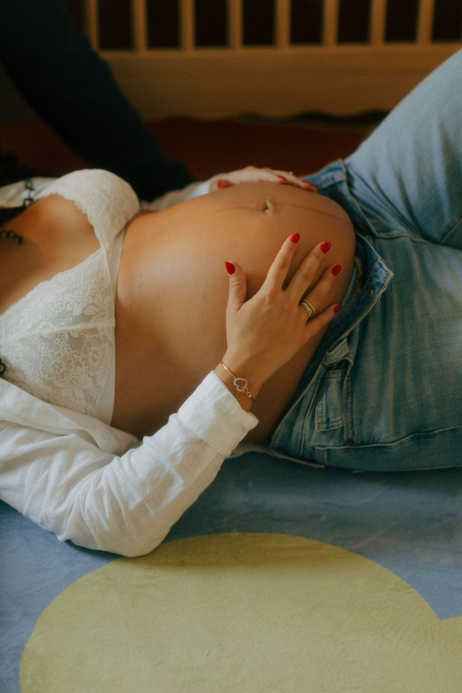 Pregnant woman lying down, cradling her bare baby bump with red nails, wearing jeans and white lace top.