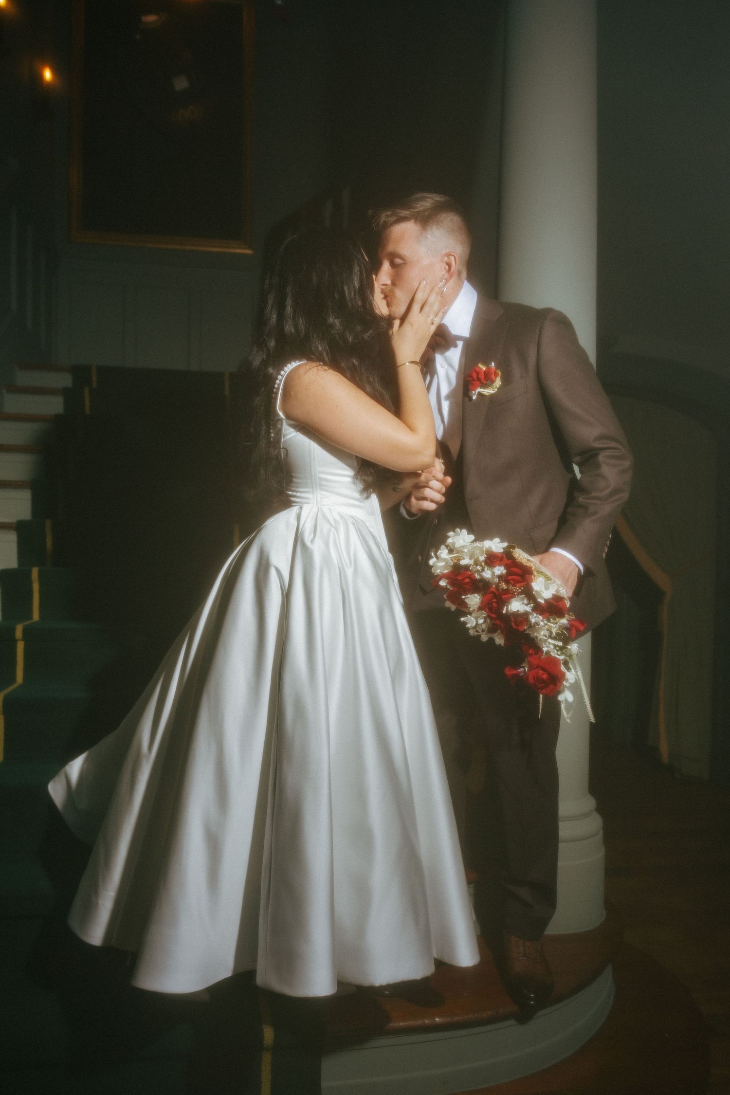 Bride and groom sharing romantic kiss near staircase column, groom holding red and white bridal bouquet.