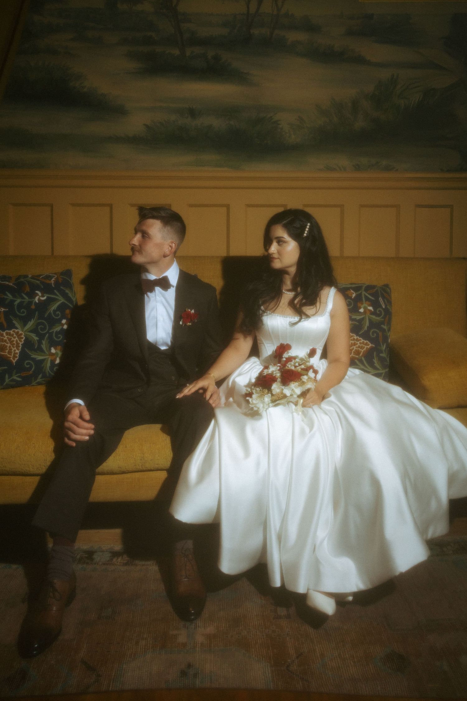 Bride in white satin gown and groom in black suit sitting together on yellow sofa with floral mural backdrop.