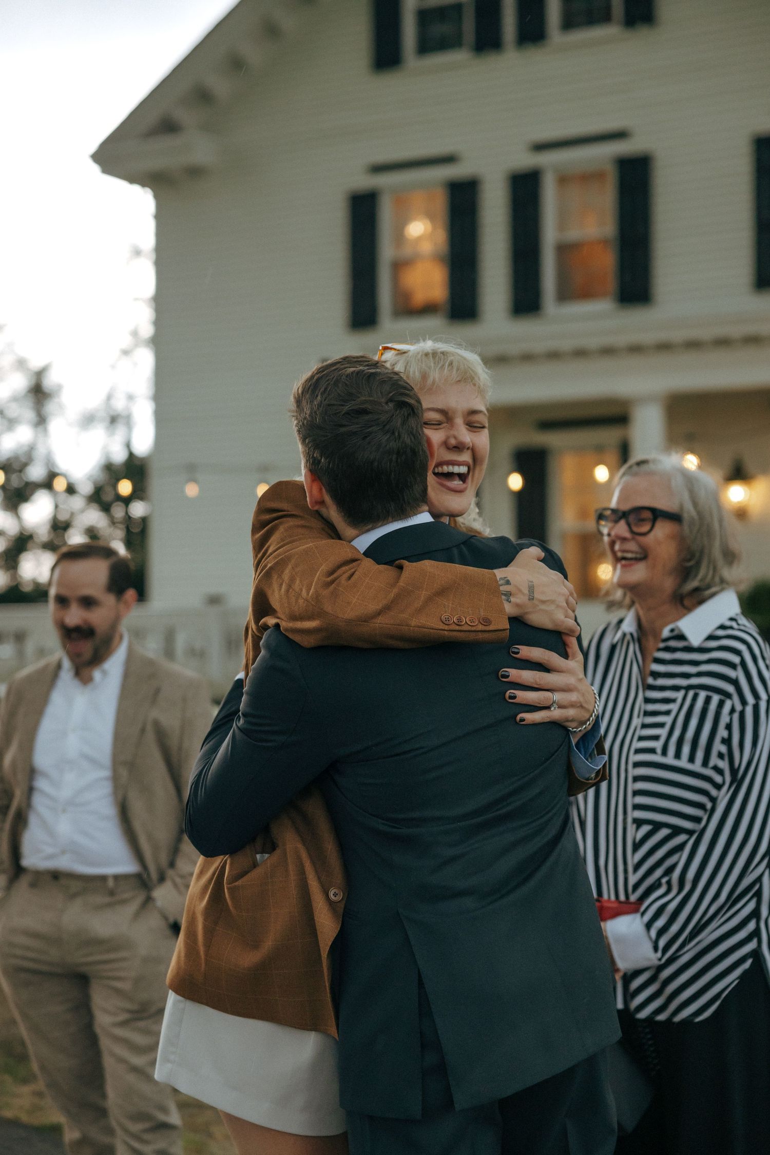 Joyful guests hugging and laughing outside a white farmhouse at a wedding reception during dusk.