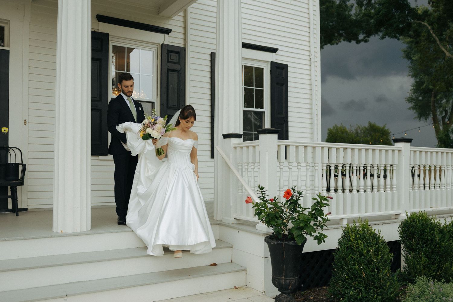 Bride and groom descend white porch steps of elegant venue, bride holding bouquet in classic ball gown wedding dress.