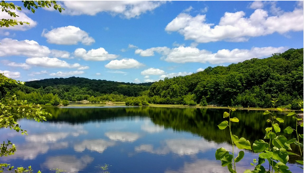Serene lake reflecting blue sky and white clouds surrounded by lush green forested hills on a sunny day.
