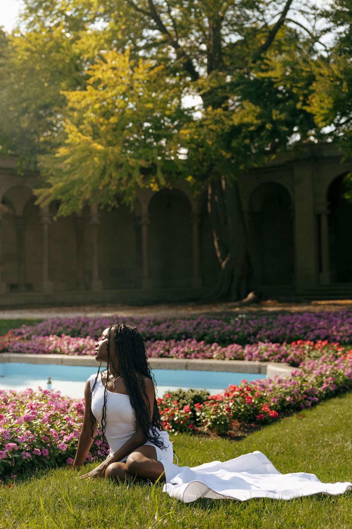 Woman in white dress sits on grass by flower garden and pool in sunlit park with arched stone building.