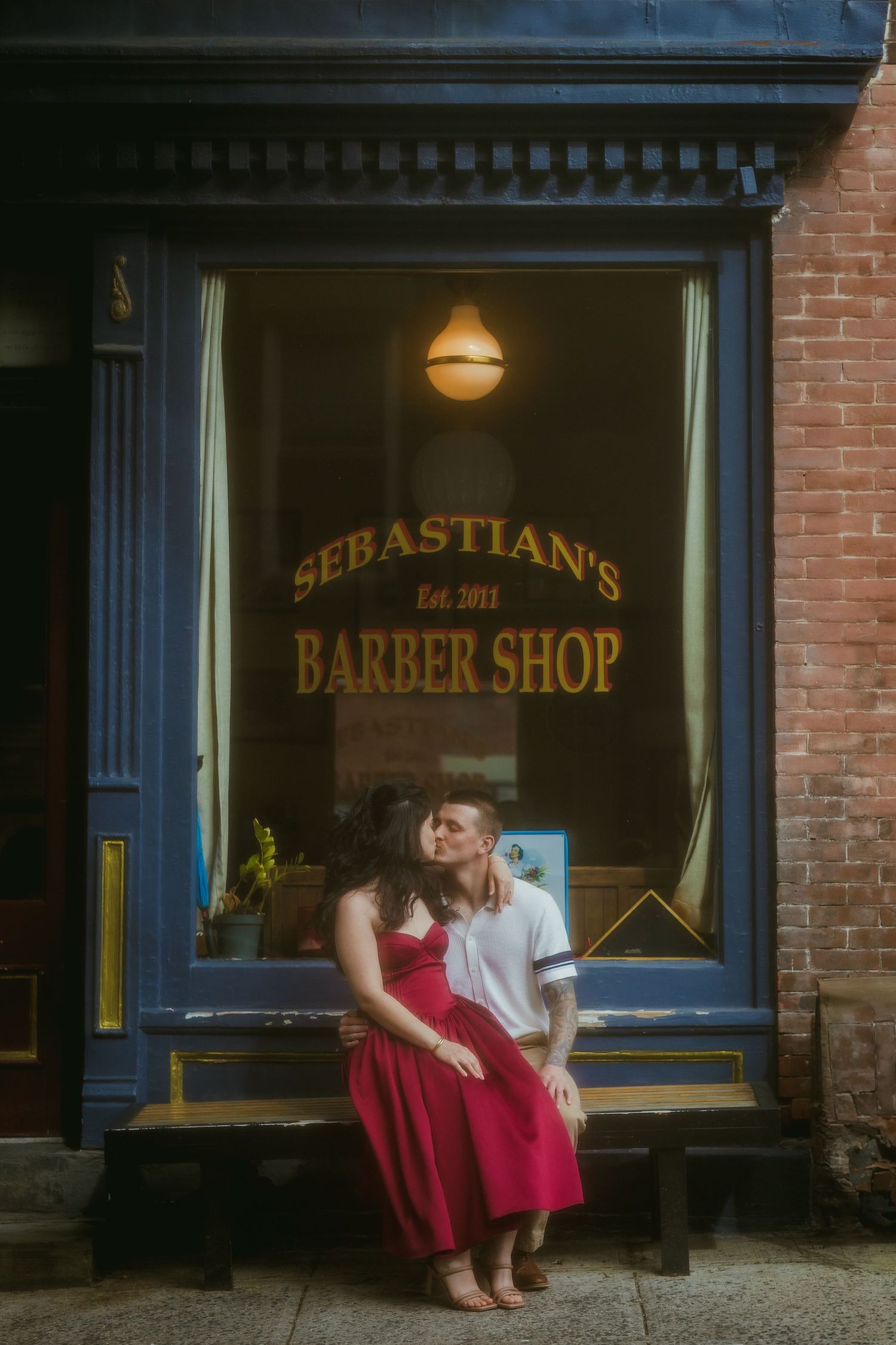 Couple sharing a romantic kiss outside Sebastian's Barber Shop, woman in red dress, urban brick storefront backdrop.