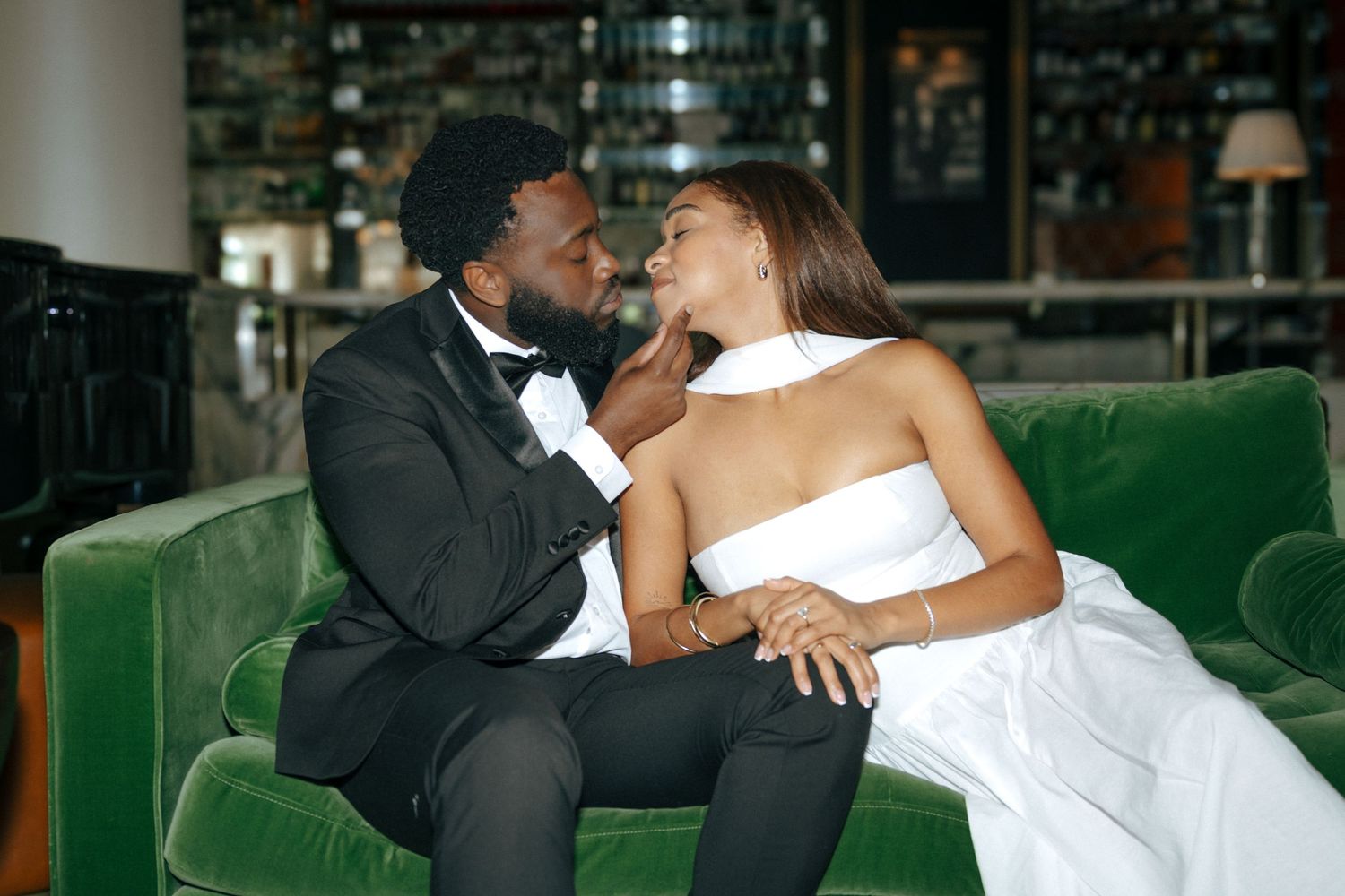 A couple in wedding attire shares a romantic moment on a green velvet sofa, the groom in a tuxedo and bride in a white gown.