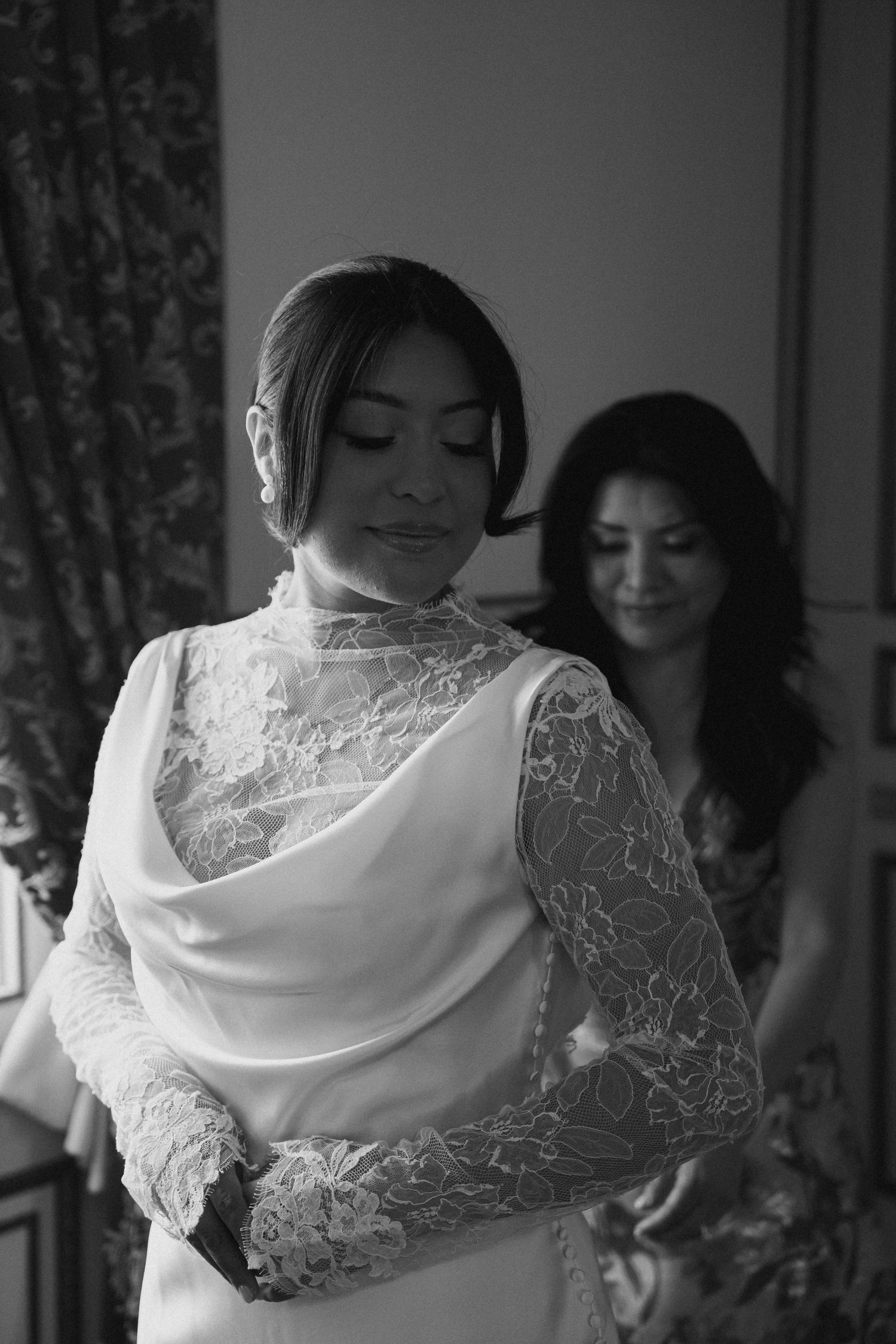 Black and white photo of a bride in lace wedding dress being helped by bridesmaid during bridal preparations.