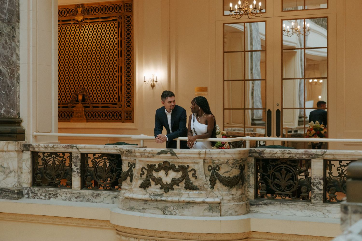 Interracial couple standing together on a marble balcony inside a grand ornate building with chandeliers.
