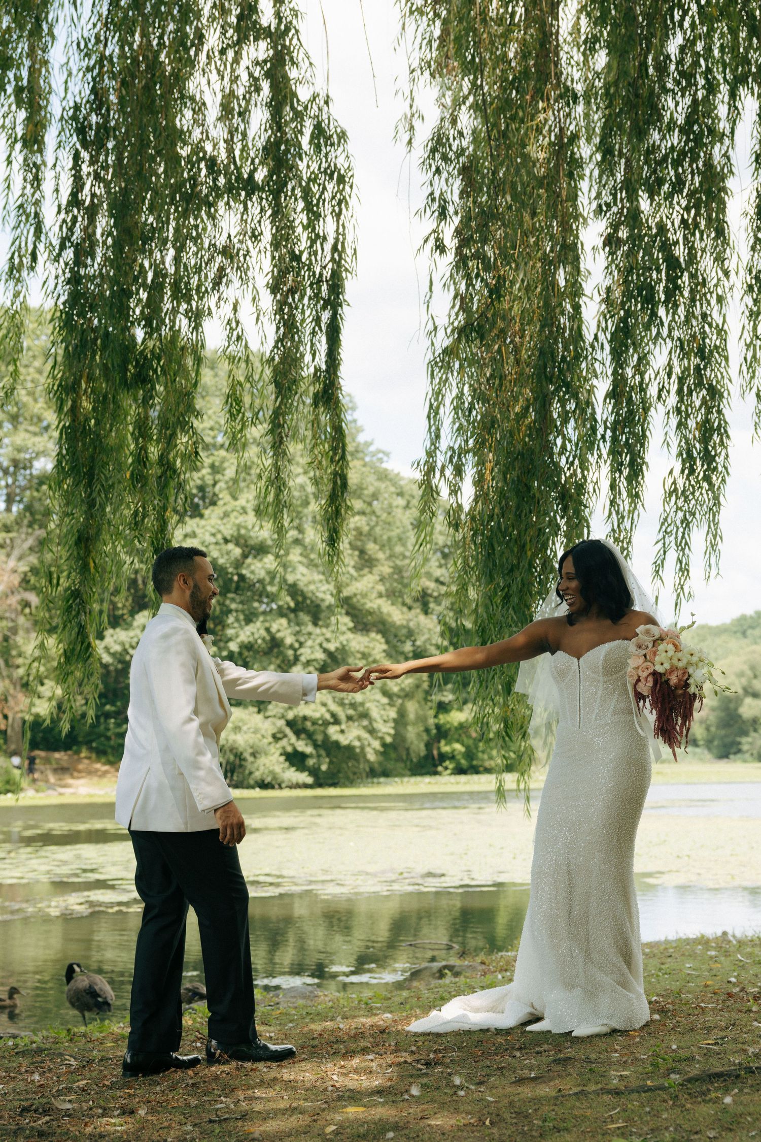 Bride and groom hold hands under weeping willow tree by tranquil lake on wedding day.
