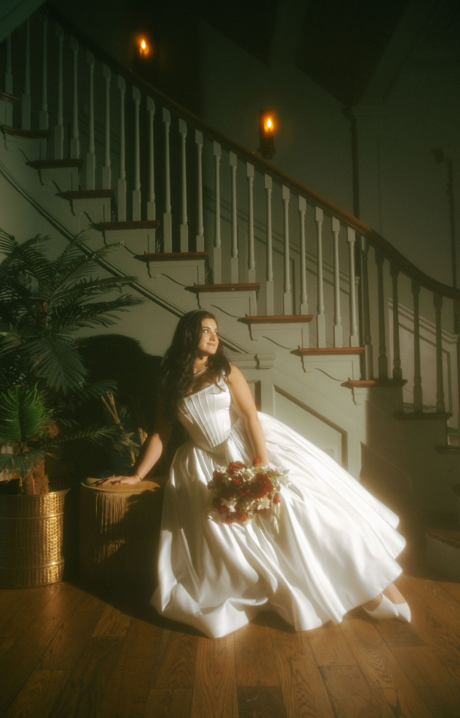 Bride in a white ball gown sitting by a staircase holding a bouquet in warm candlelit indoor lighting.