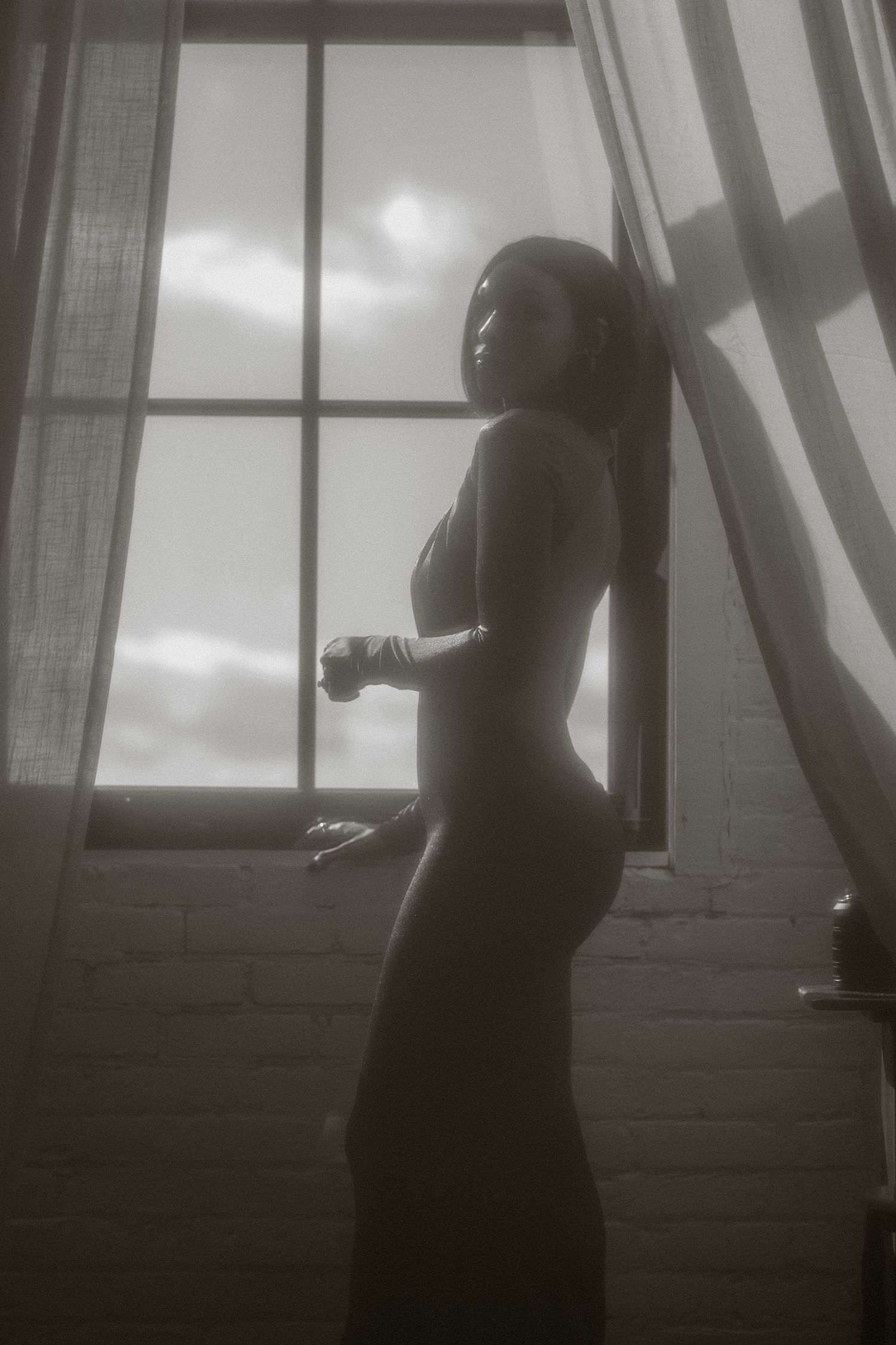 Black and white silhouette of woman in lingerie standing by large window with sheer curtains.
