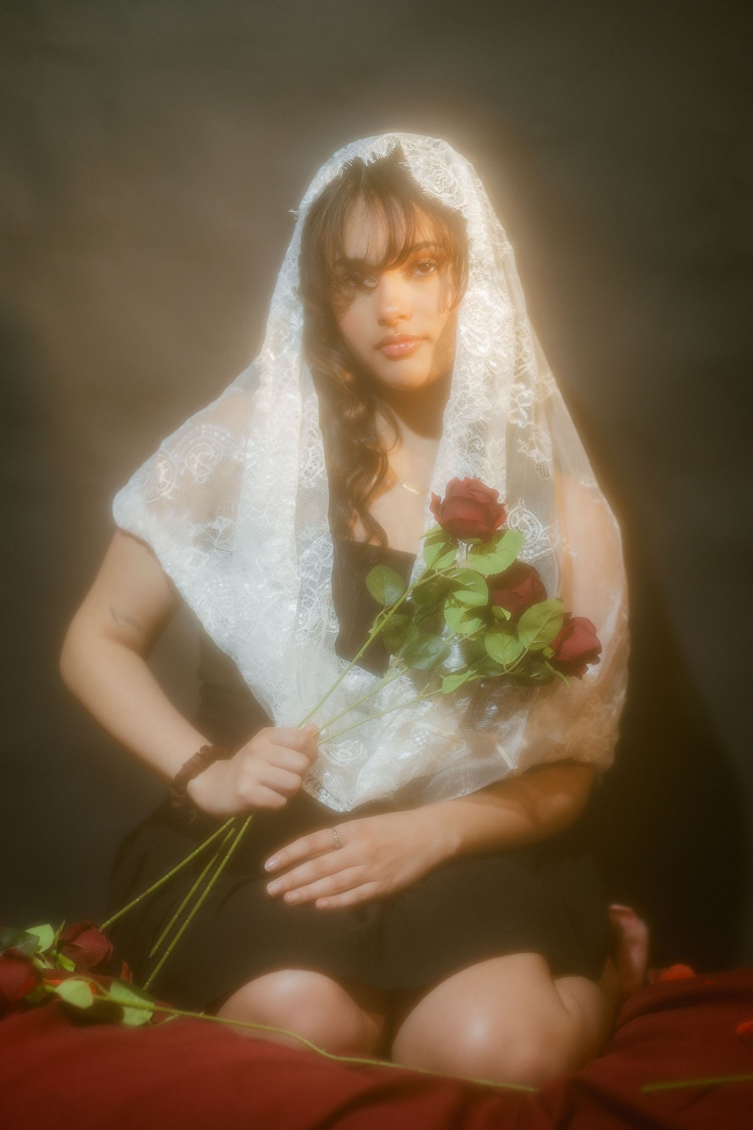 Woman wearing white lace veil holding red roses on dark dramatic background.