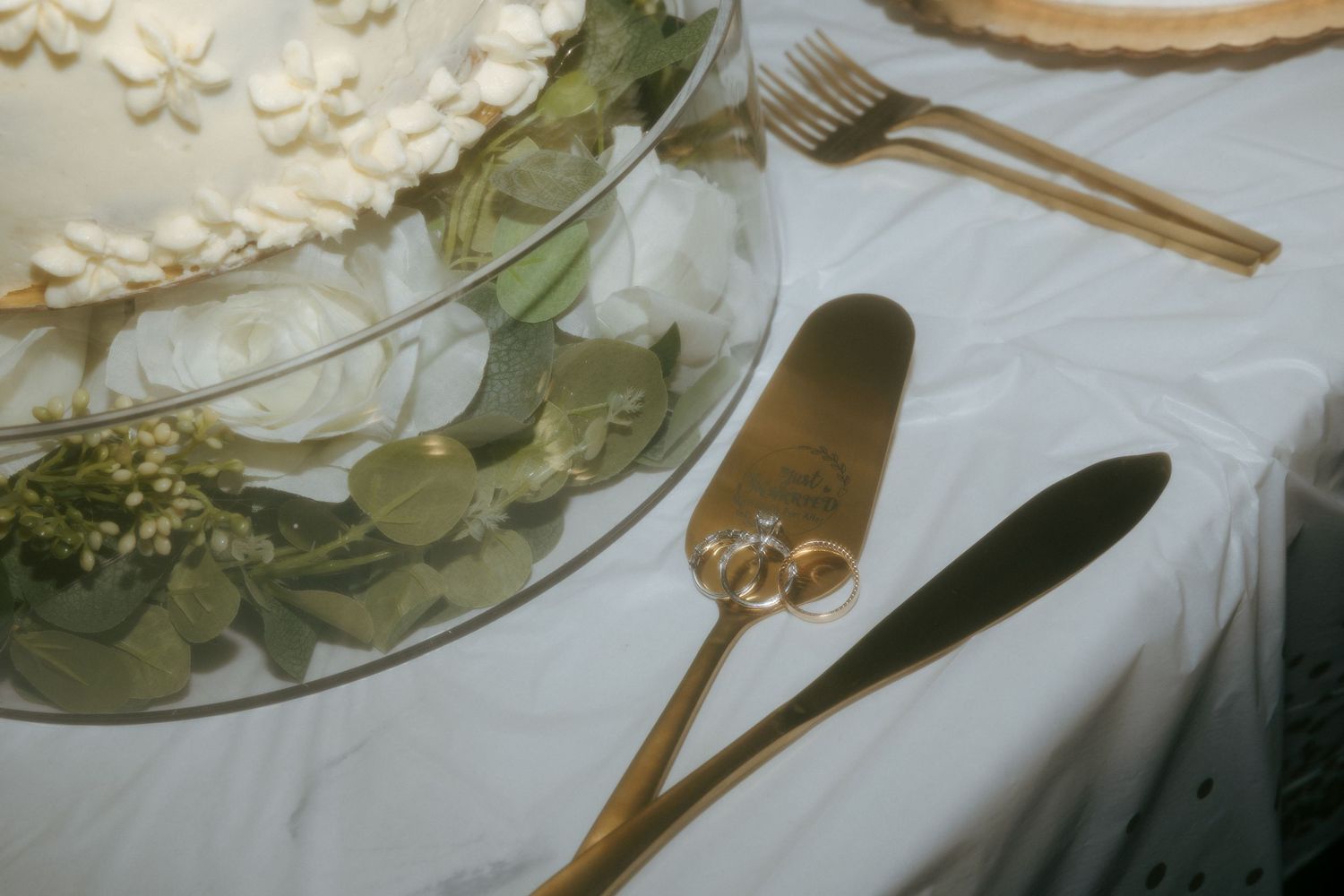 Gold wedding cake server and knife set with rings detail beside white frosted wedding cake on elegant white tablecloth.