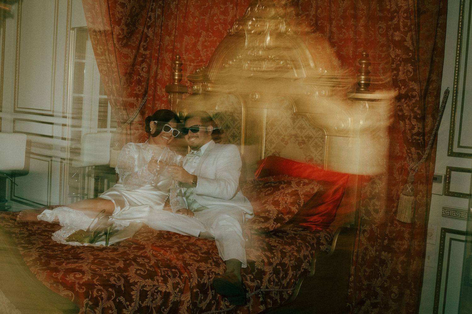 Two people in white outfits sit together on an ornate red bed in an elegant room with draped curtains.