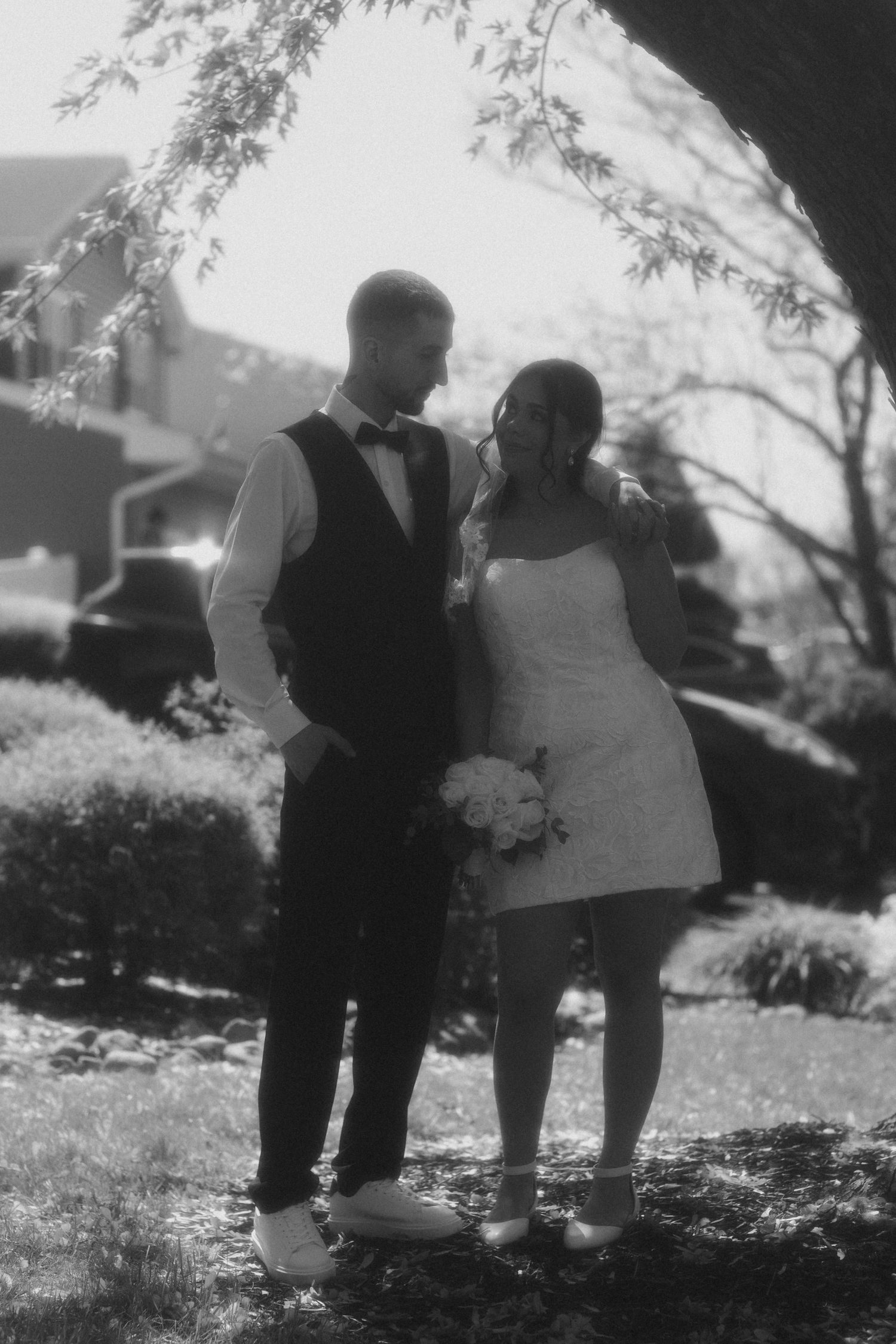 Black and white photo of a bride in short white dress holding bouquet standing with groom in vest and bow tie outdoors.