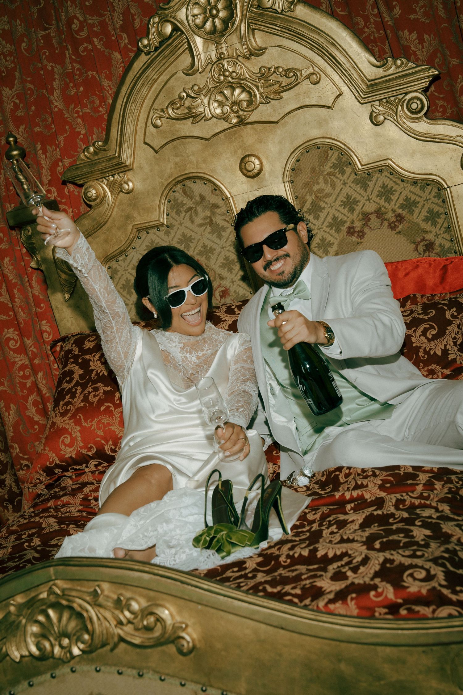 Bride and groom in sunglasses celebrating with champagne bottles on ornate gold bed with red velvet decor.
