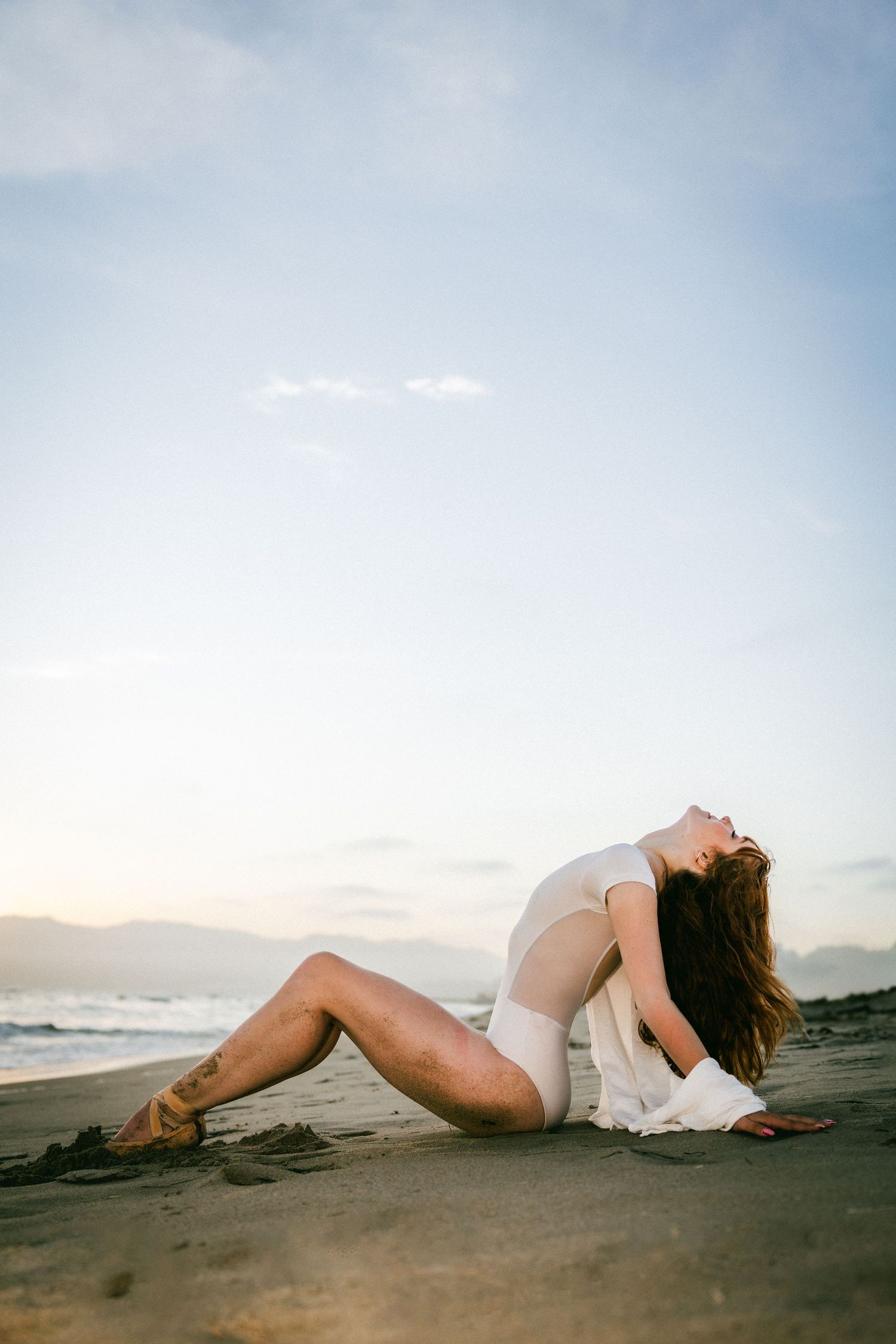 Woman in white bodysuit posing dramatically on sandy beach at sunset with ocean waves in background.