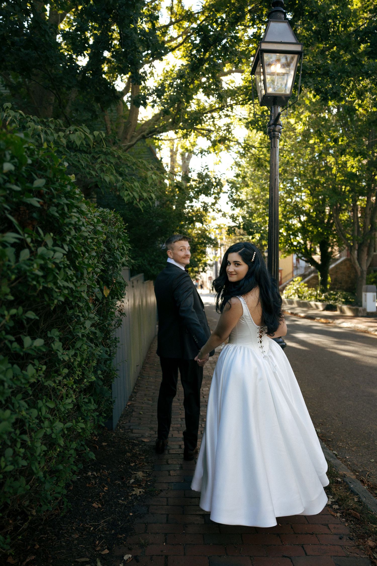 Bride in white gown and groom in black suit hold hands walking along tree-lined brick pathway.