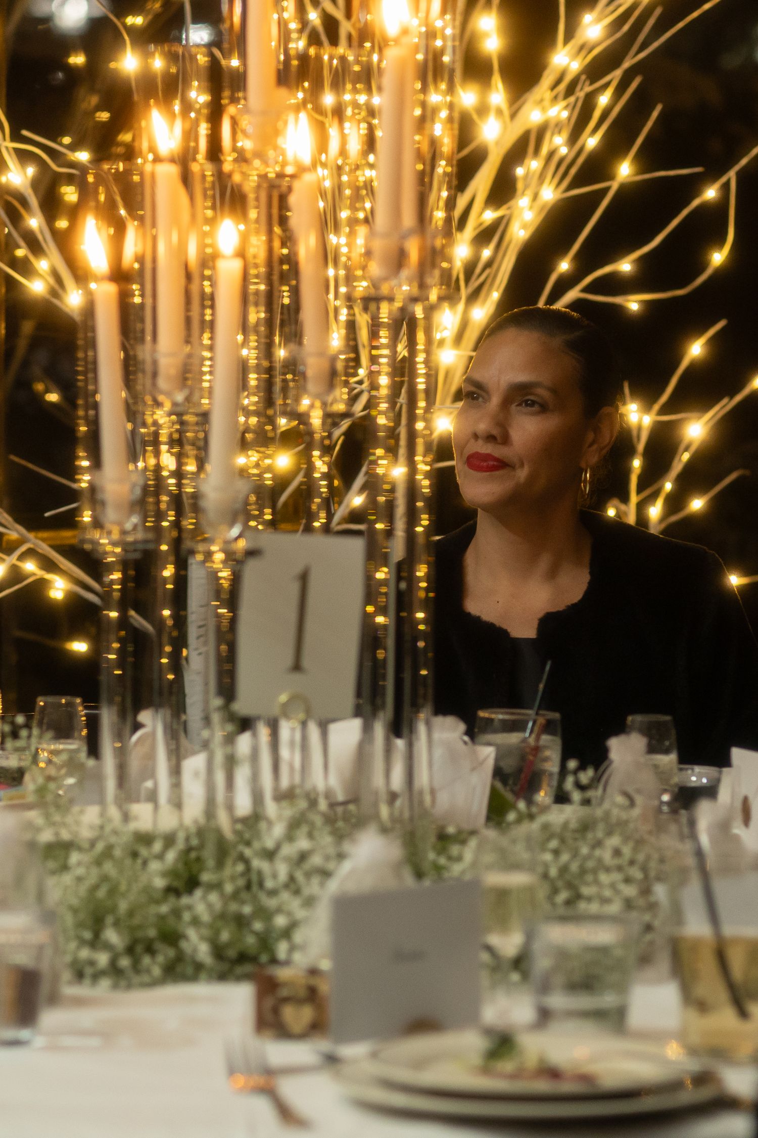 Woman in black seated at elegantly decorated wedding table with candelabras and warm fairy lights.