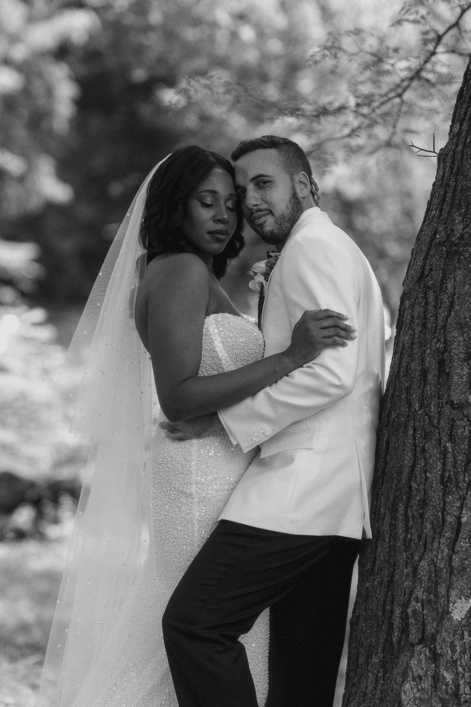 Black and white portrait of bride and groom embracing romantically against a tree.