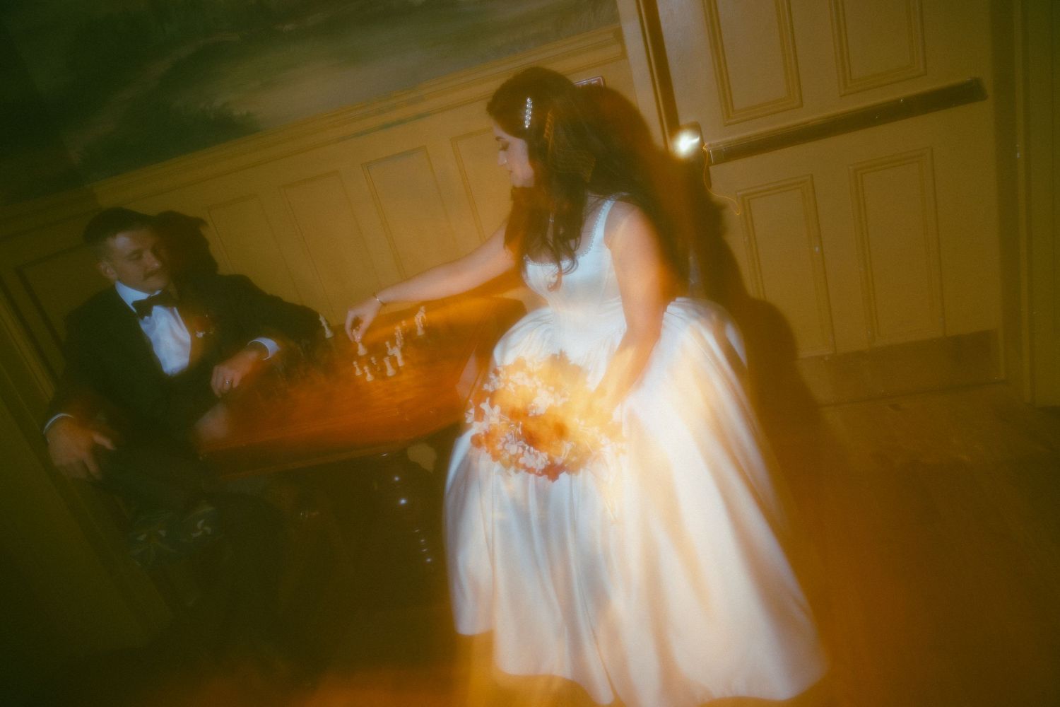 Blurry artistic wedding photo of bride in white gown holding bouquet reaching toward groom in dimly lit room.