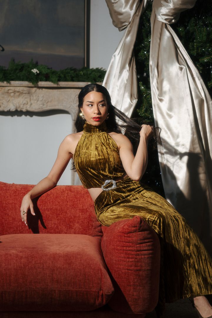 Elegant woman in gold velvet halter dress seated on red sofa near draped satin and holiday greenery.