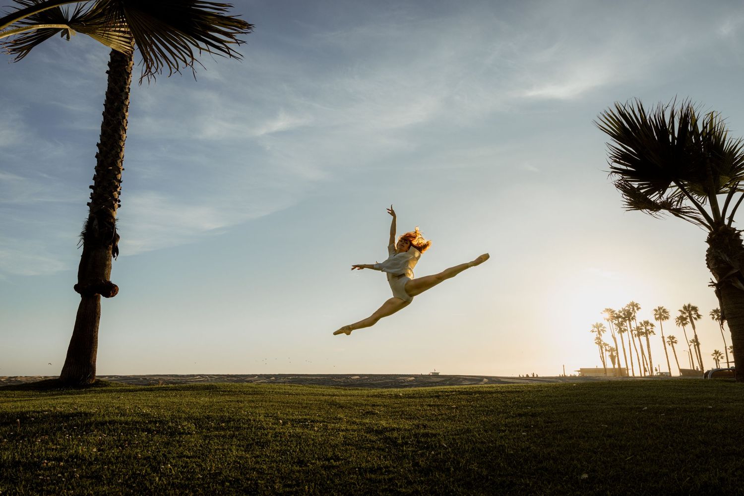 Woman performing a grand jete leap between palm trees at sunset on green lawn.