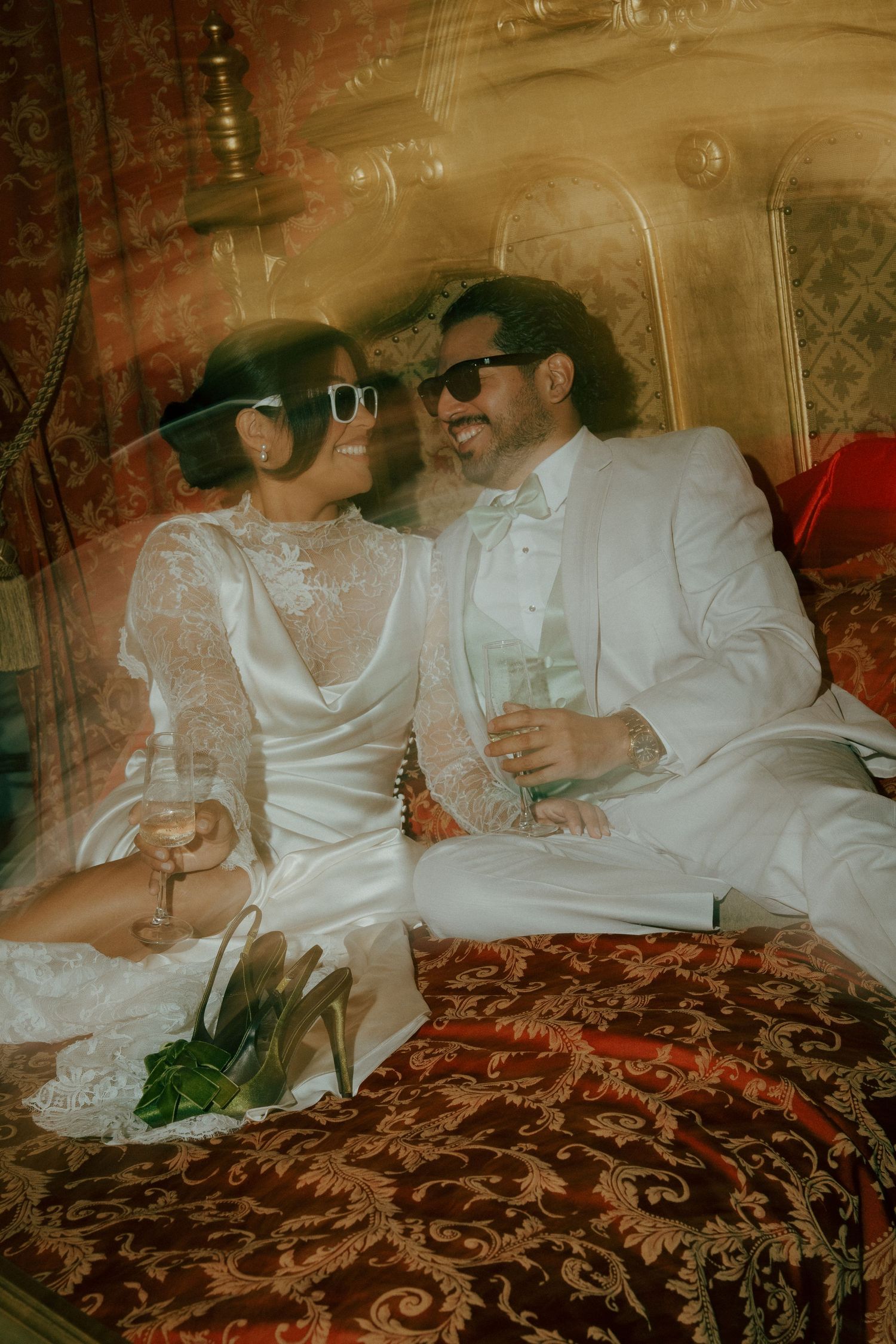 Newlyweds in white wedding attire and sunglasses share champagne on ornate red velvet furniture.