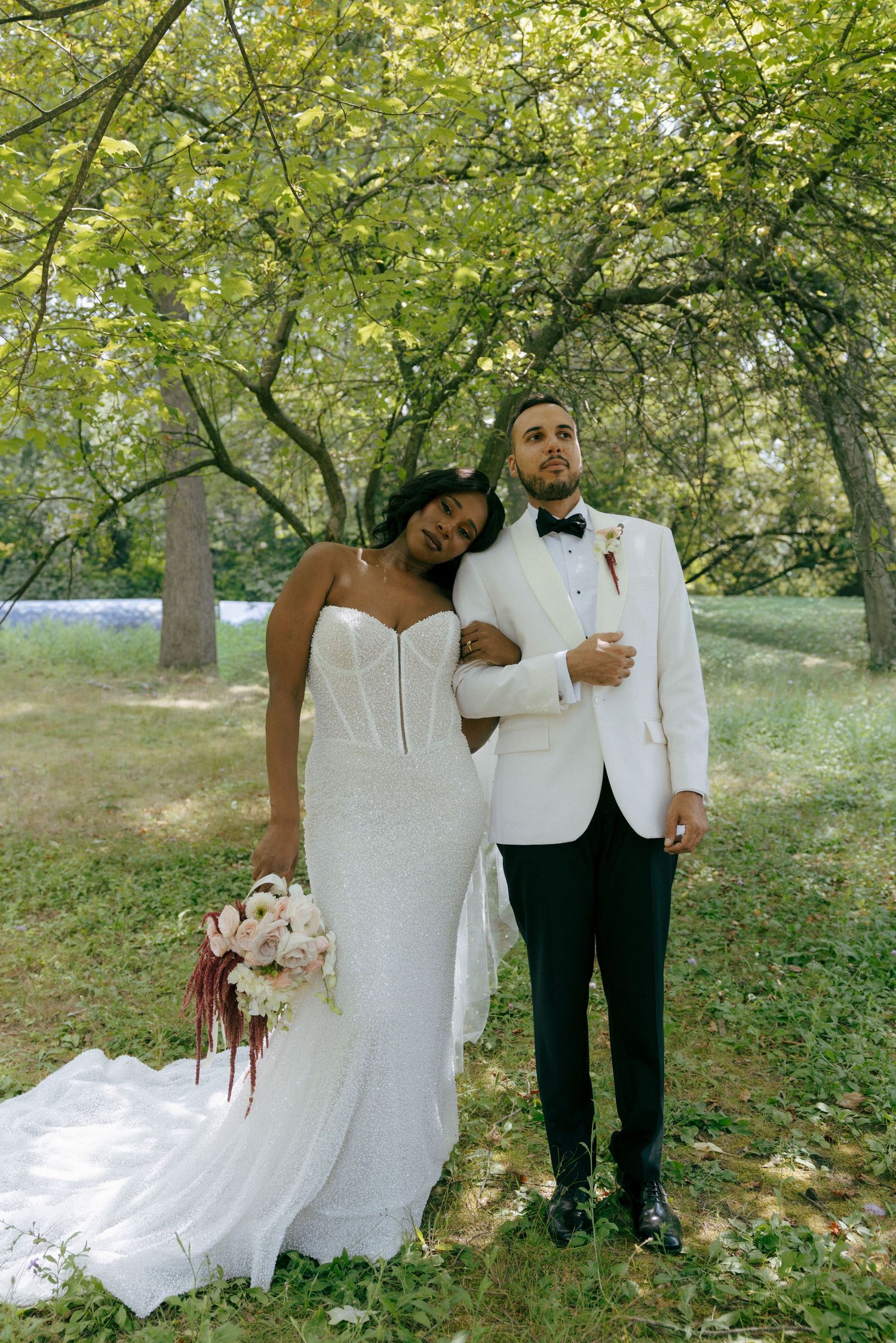 Interracial couple in wedding attire embracing under green trees outdoors.