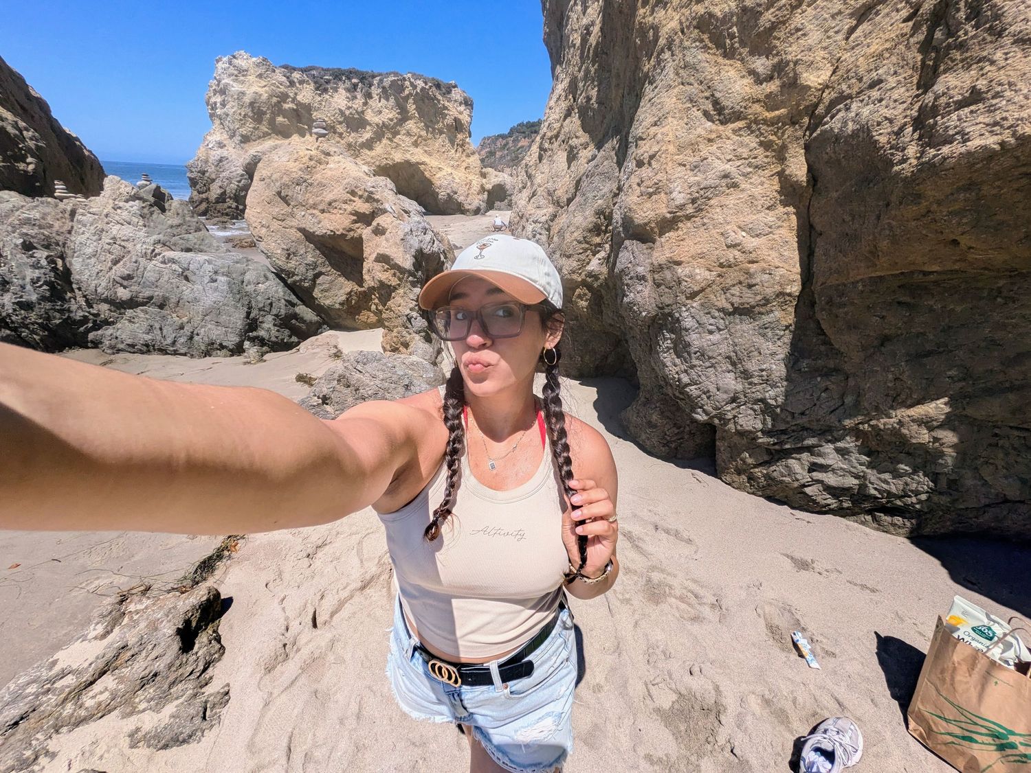 Woman taking a selfie on a sandy beach with large rock formations and blue sky in the background.