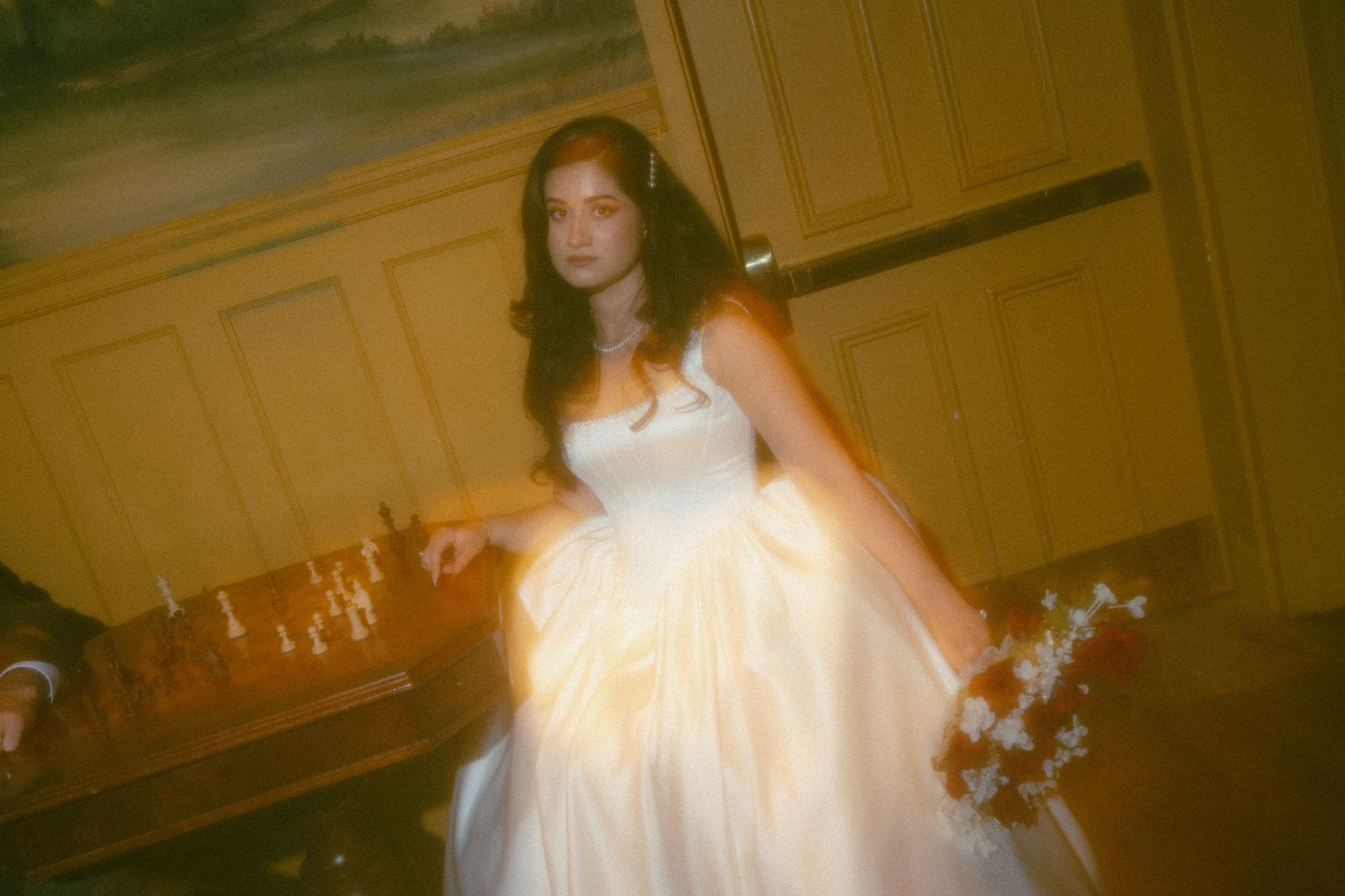 A bride in a white ballgown holds a floral bouquet in a warmly lit vintage room.