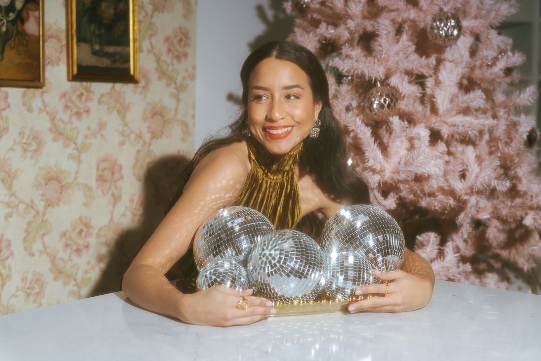 Smiling woman in gold halter top embracing silver disco balls on table near pink Christmas tree.