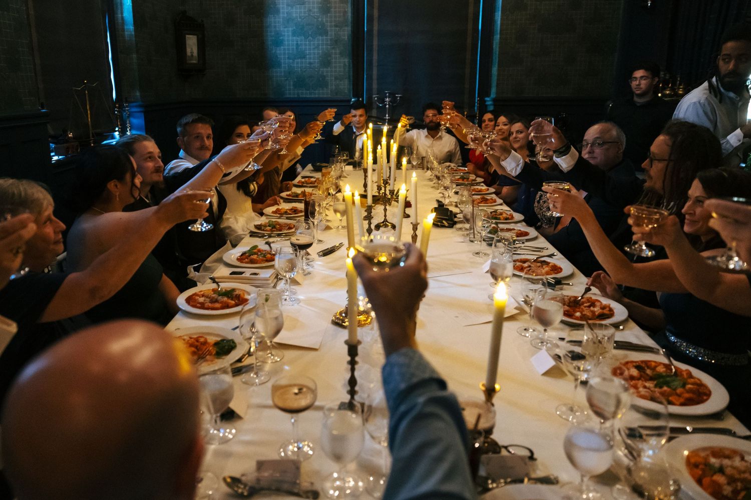 Wedding guests raising glasses in candlelit toast over long dinner table with elegant place settings.