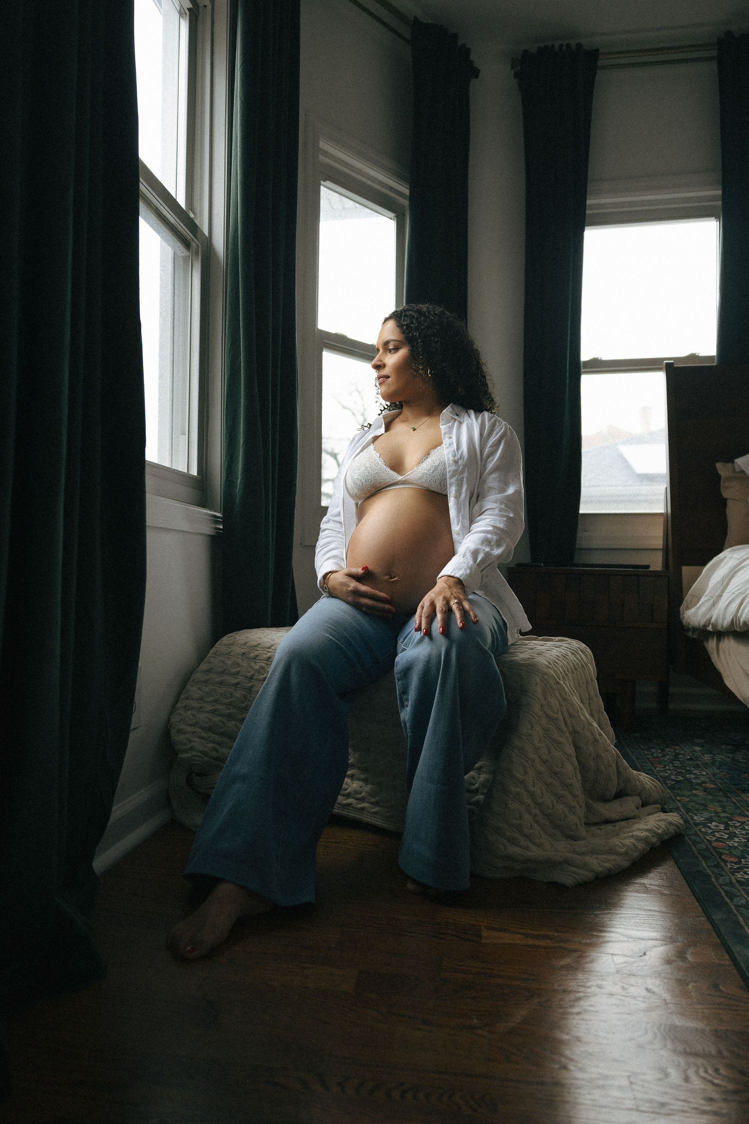 Pregnant woman in white bra and open shirt sits on bed by window, cradling her baby bump in a cozy bedroom.